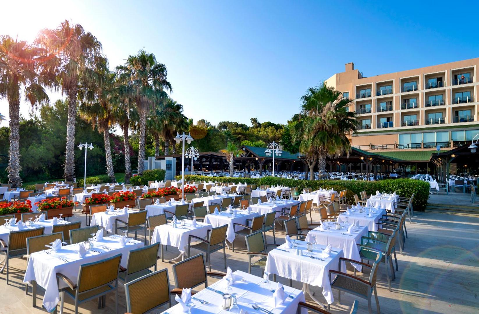 Turquoise Hotel āra restorāna terase ar balti klātiem galdiem, palmām un skatu uz viesnīcas ēku saulainā dienā Sidē, Turcijā.