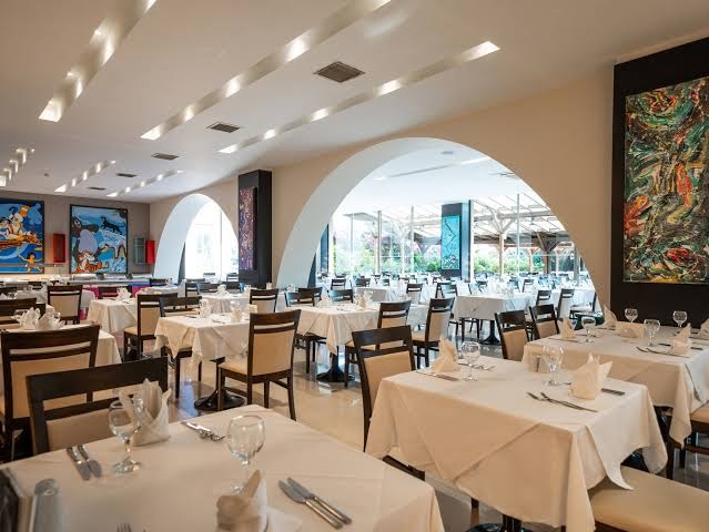 Turquoise Hotel iekštelpu restorāns ar elegantiem balti klātiem galdiem, arkām un mākslas darbiem pie sienām.