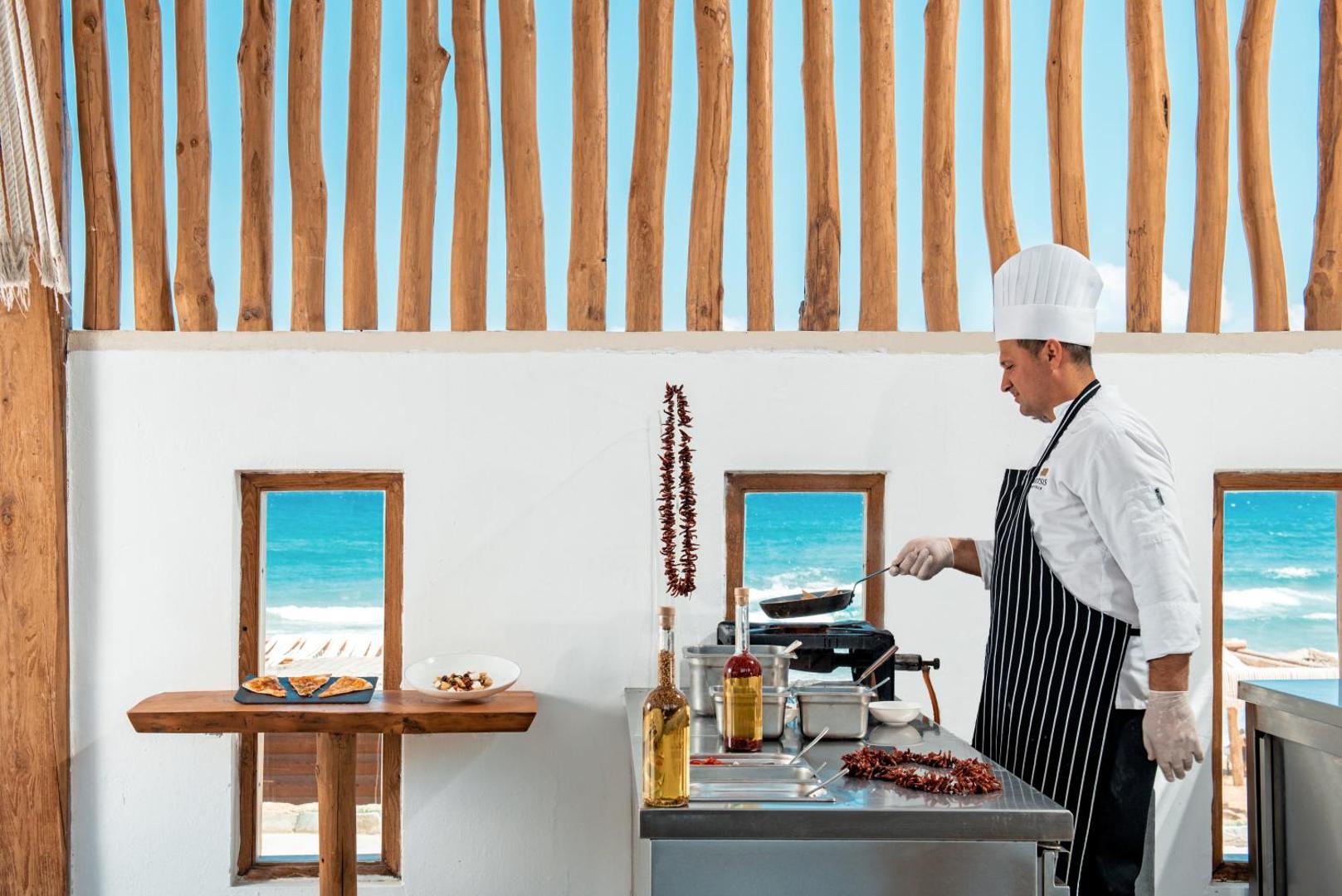 Mitsis Rinela Beach Resort šefpavārs gatavo ēdienu atvērtā tipa virtuvē ar skatu uz jūru — svaigas sastāvdaļas un servēšana