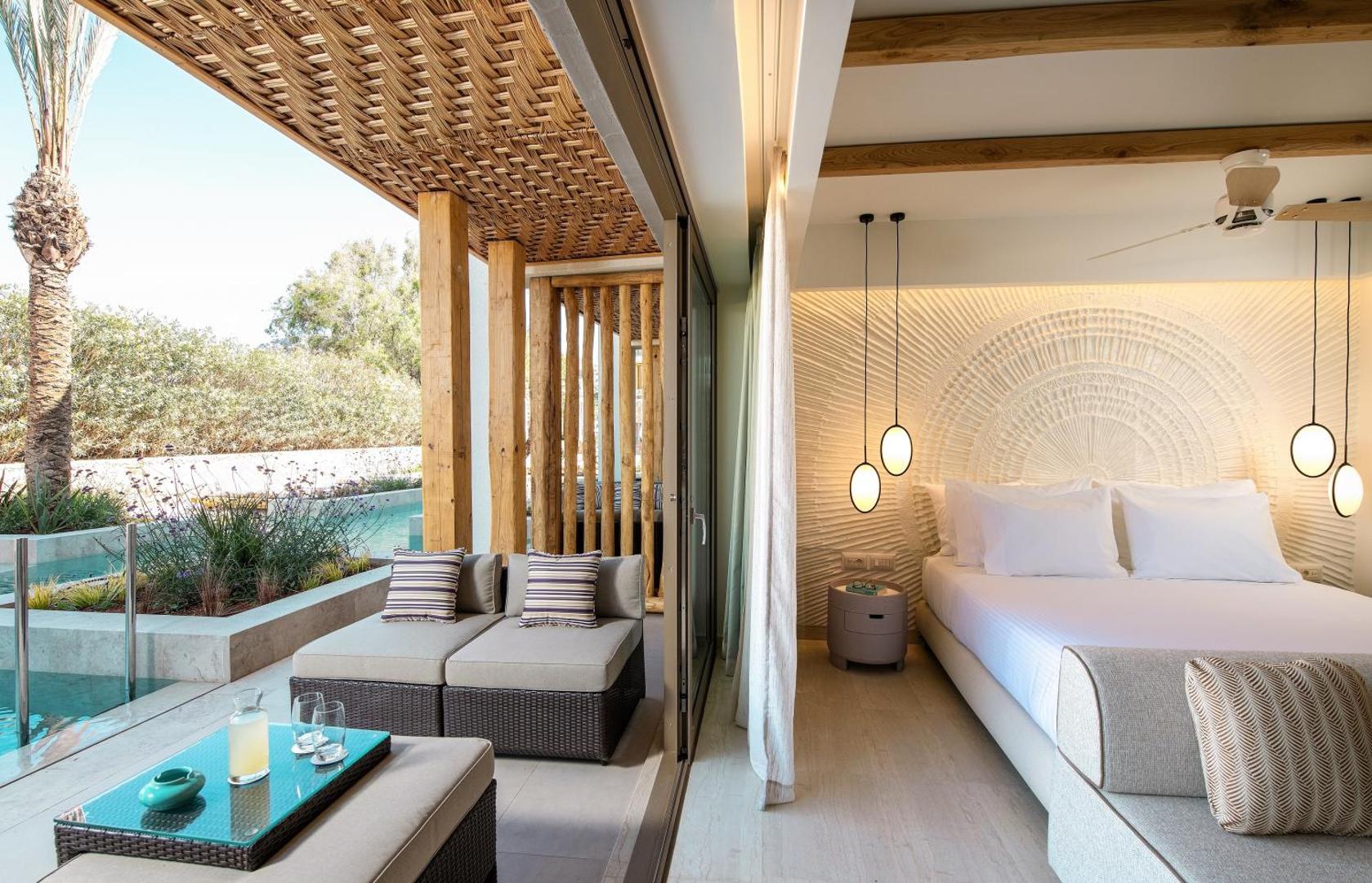 Mitsis Rinela Beach Resort numurs ar atvērtu pāreju uz terasi, kur atrodas atpūtas krēsli un baseins piekrastes zonā