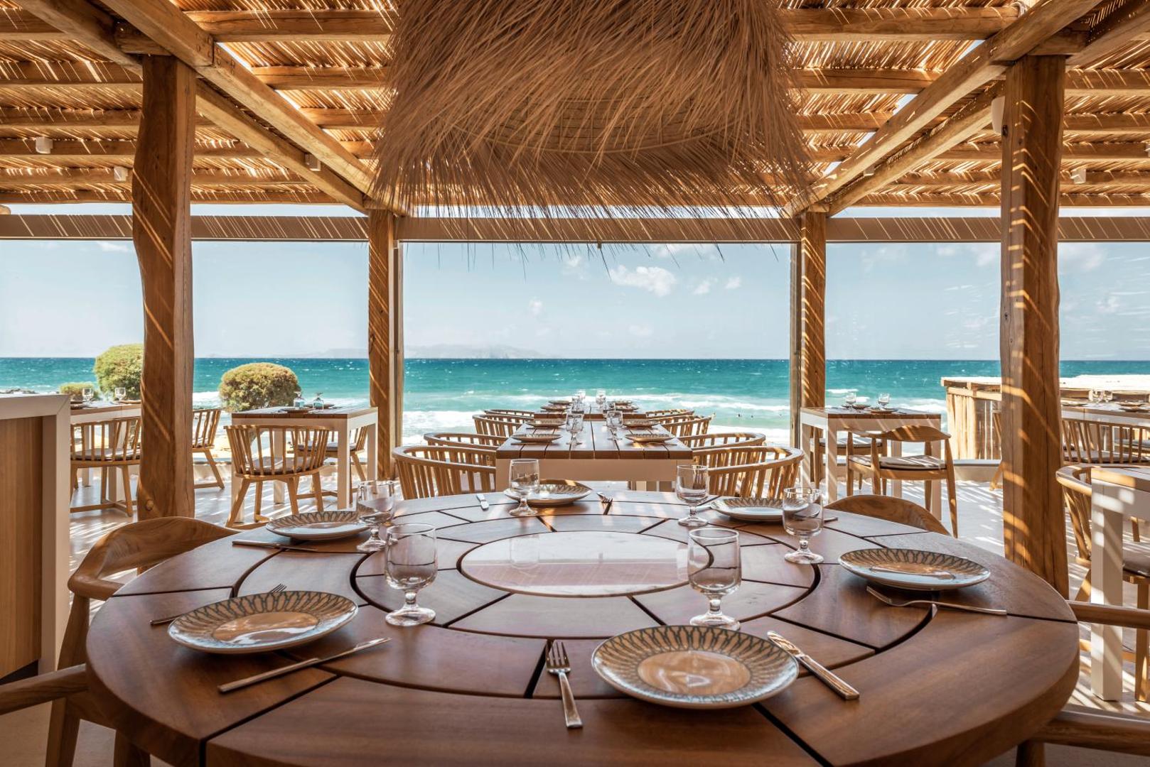 Mitsis Rinela Beach Resort restorāna apaļais galds ar skatu uz tirkīzzilo jūru — elegants galda klājums zem niedru jumta