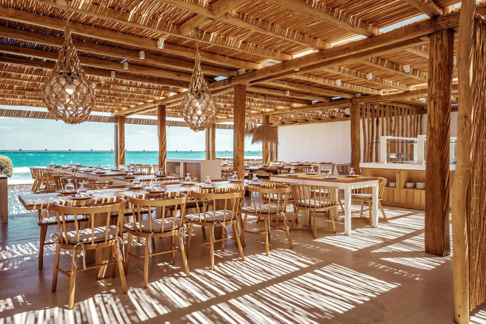 Mitsis Rinela Beach Resort restorāns ar koka interjeru un skatu uz jūru Krētā — gaiša terase ar klātiem galdiem