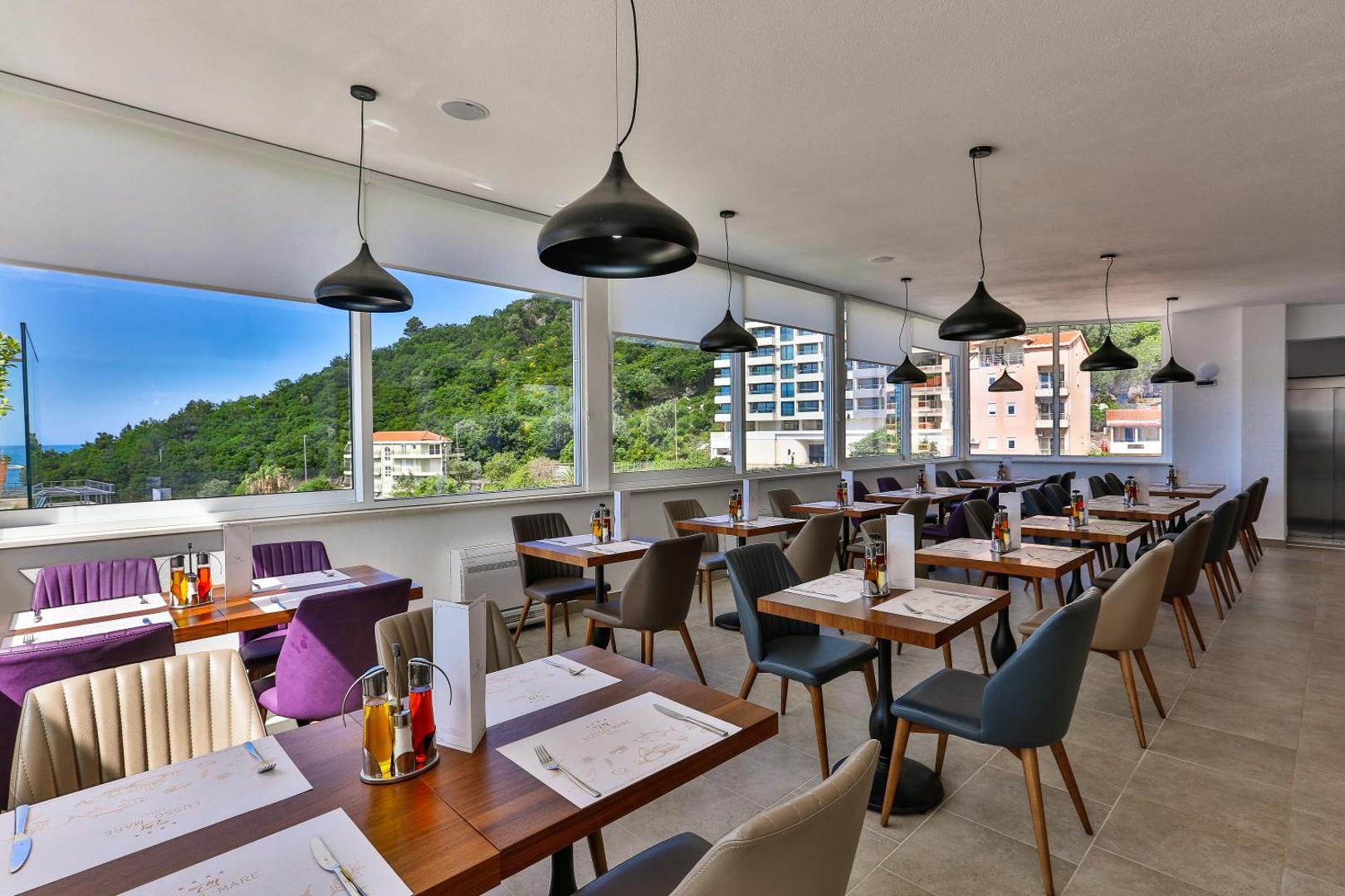 Hotel Lusso Mare restorāna iekštelpas ar modernu dizainu, koka galdiem un lieliem panorāmas logiem, pa kuriem redzama pilsēta un kalni
