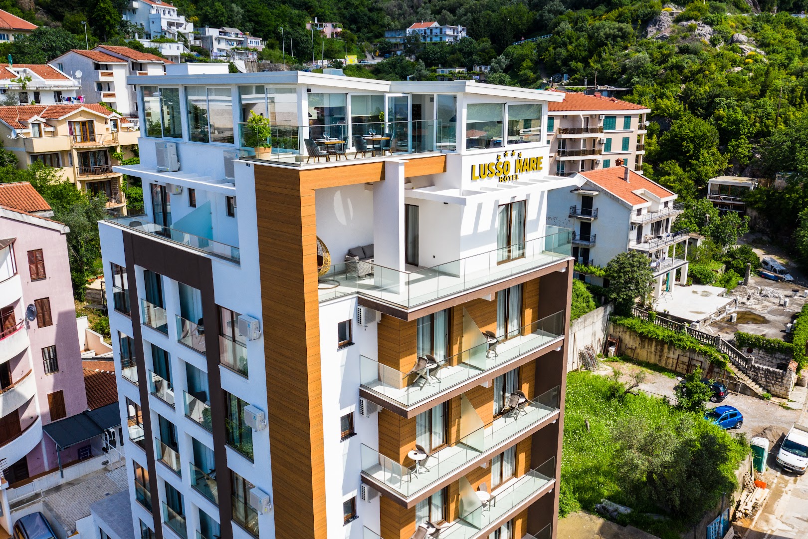 Hotel Lusso Mare fasādes tuvplāns ar stiklotiem balkoniem un jumta restorāna zonu saulainā dienā Budvas kūrortā