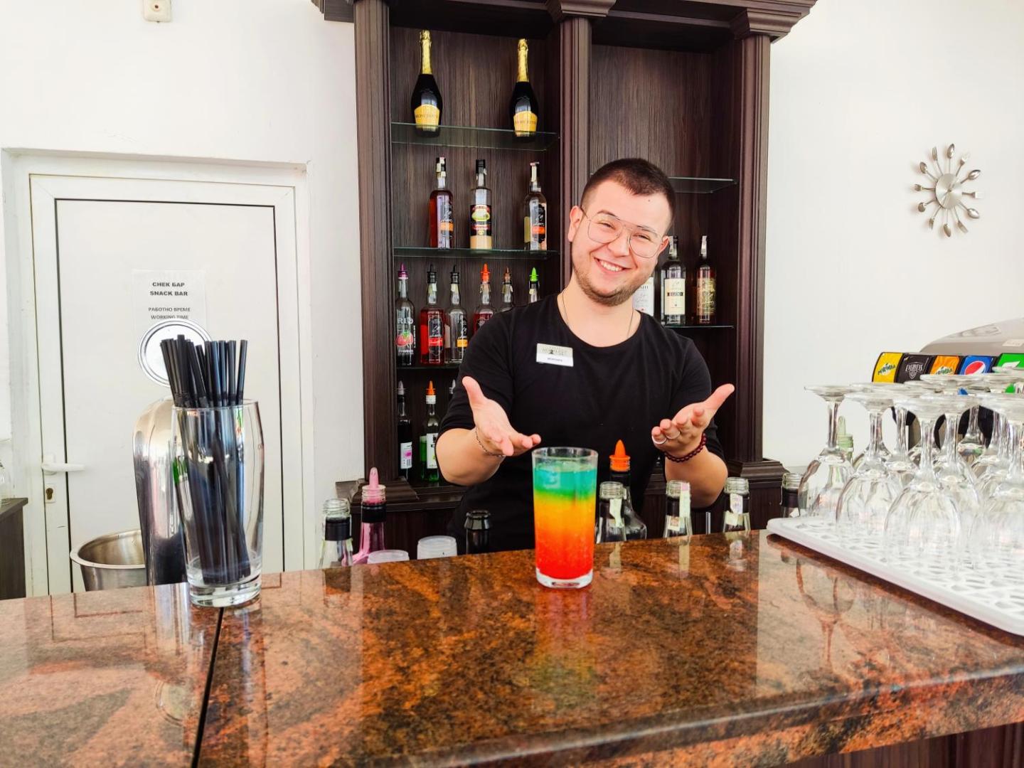 Moonlight Hotel bārmenis pasniedz krāsainu slāņaino kokteili pie marmora bāra letes — fonā dzērienu plaukti un smaidošs personāls