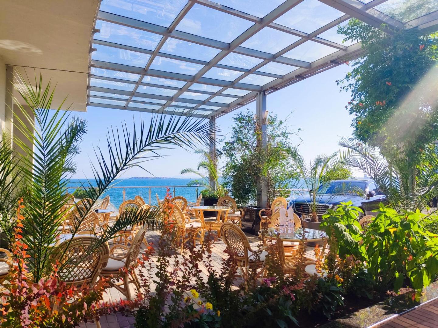 Moonlight Hotel āra terase ar pītiem krēsliem zem stikla jumta, ko ieskauj zaļumi un skats uz jūru