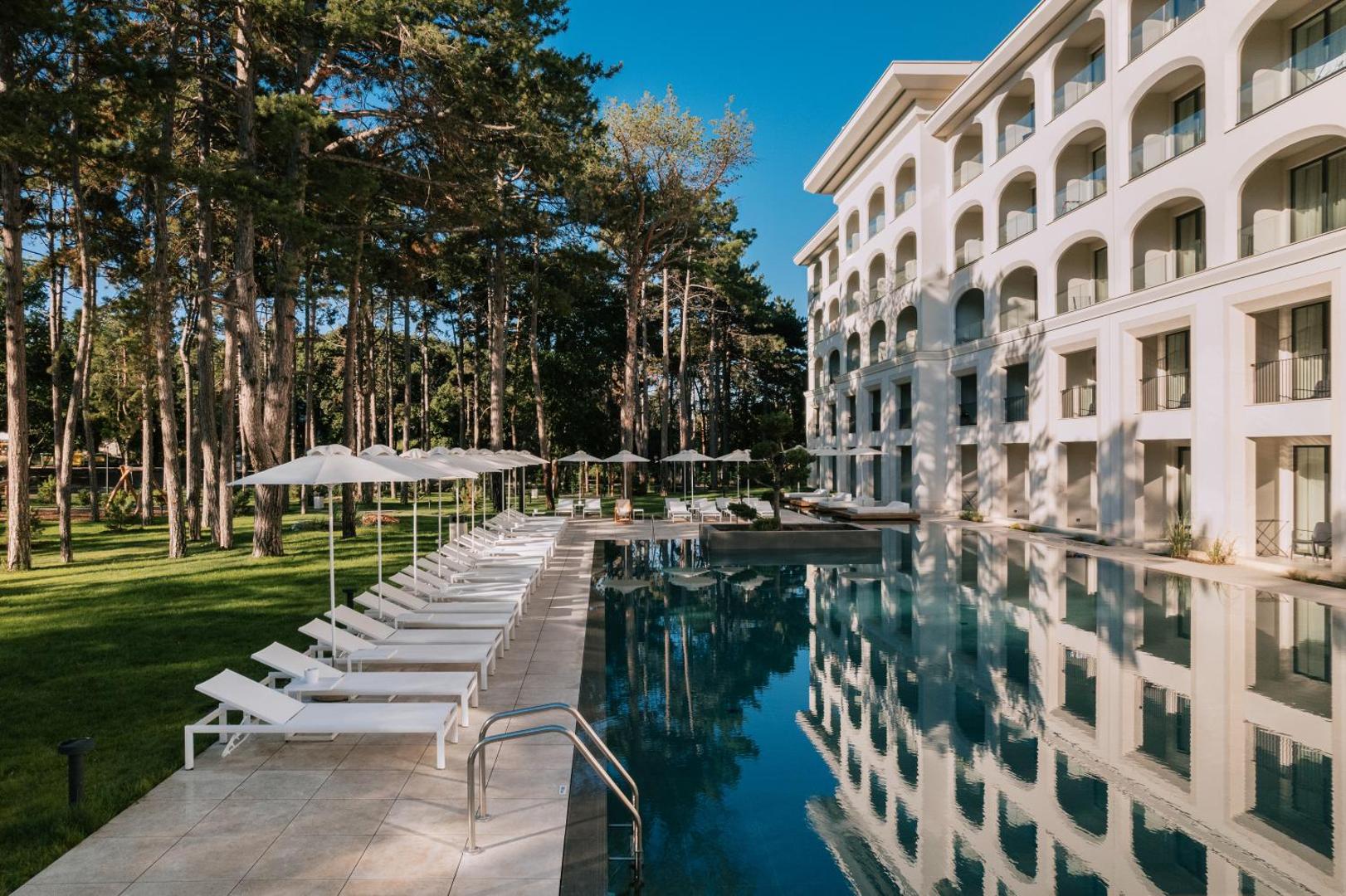 Aquahouse Hotel & SPA modernā viesnīcas ēka ar arkveida balkoniem blakus āra baseinam un augstām priedēm saulainā dienā