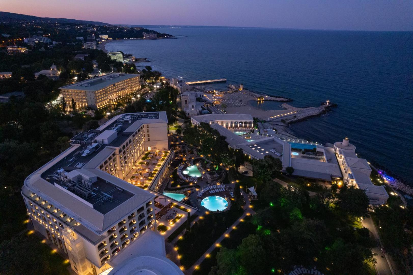 Aerofoto skats uz Aquahouse Hotel & SPA kompleksu krēslas stundā pie Melnās jūras — izgaismoti baseini, dārzi un piekrastes līnija