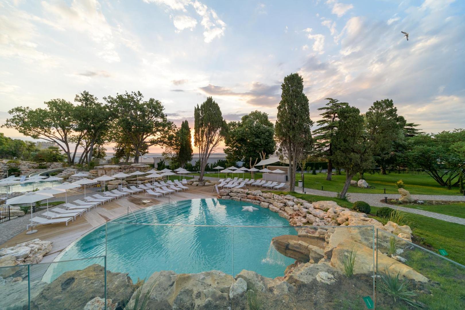 Aquahouse Hotel & SPA āra baseins ar akmens elementiem un ūdenskritumu saulrieta laikā, ko ieskauj zaļš parks un atpūtas krēsli
