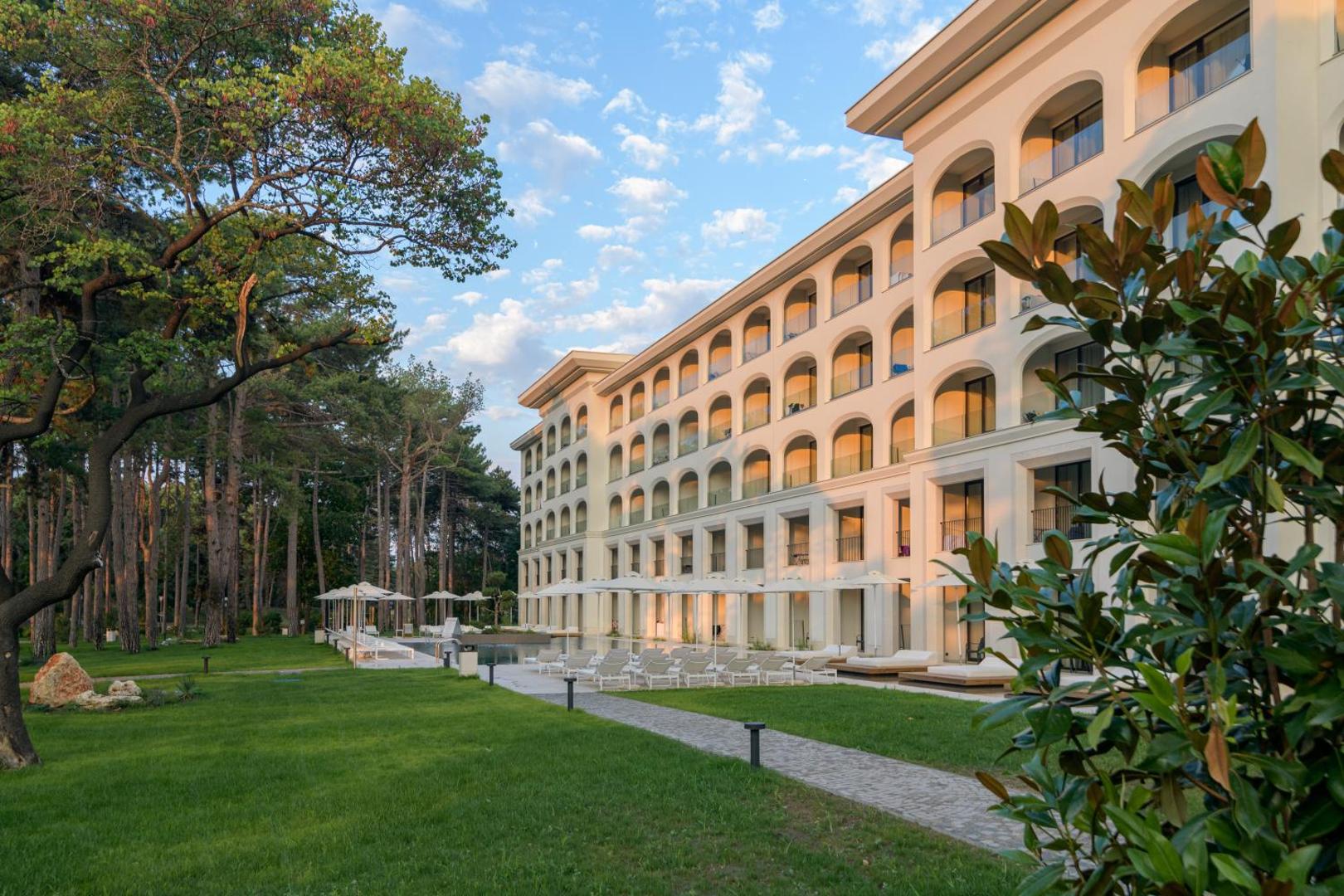 Aquahouse Hotel & SPA modernā fasāde ar arkveida balkoniem blakus priežu mežam — sauļošanās krēsli un zaļš zāliens pie numuriem