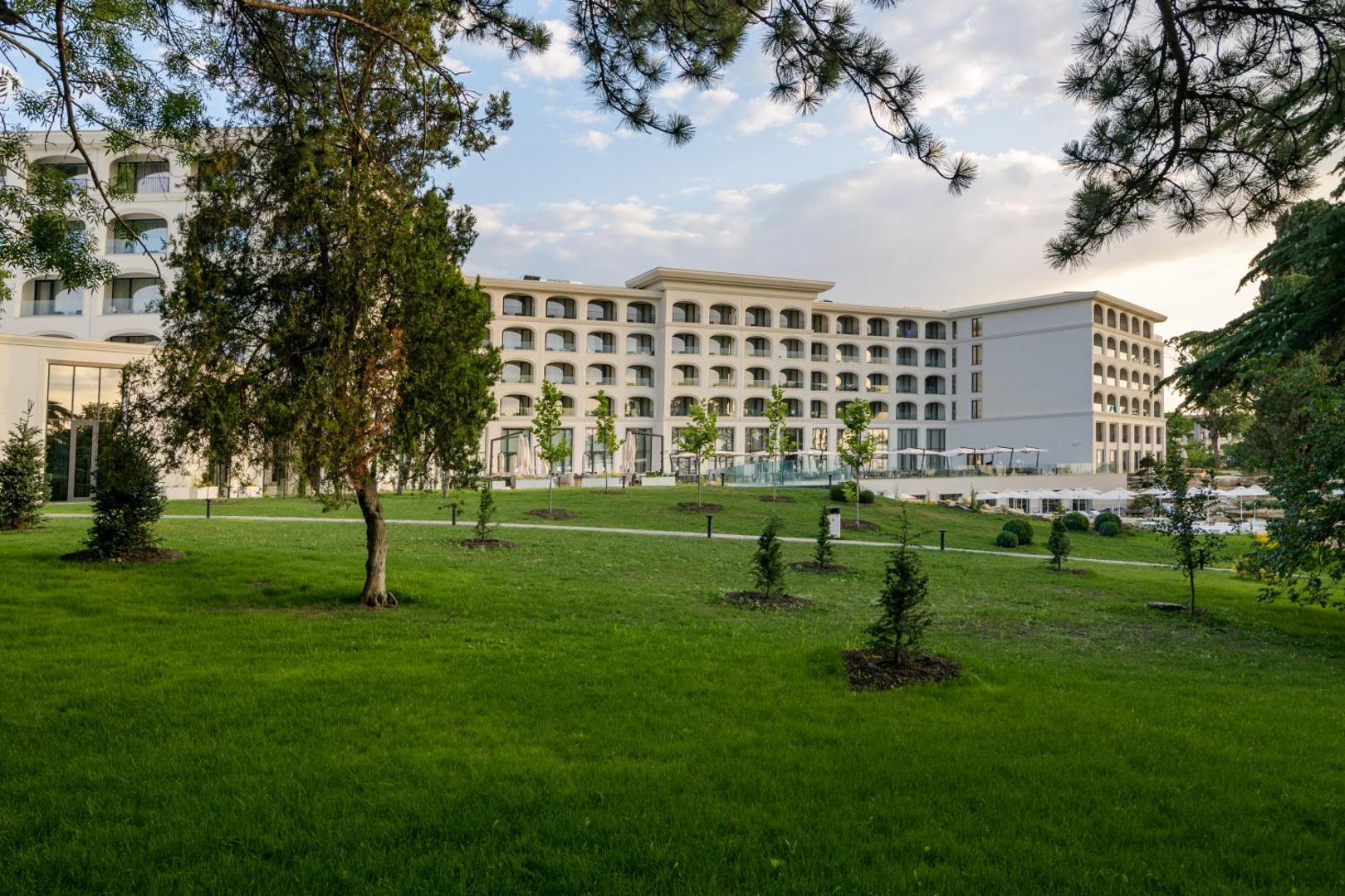 Skats uz Aquahouse Hotel & SPA balto ēku no labiekārtota dārza puses — plaši zālieni, jauni stādījumi un gājēju celiņi