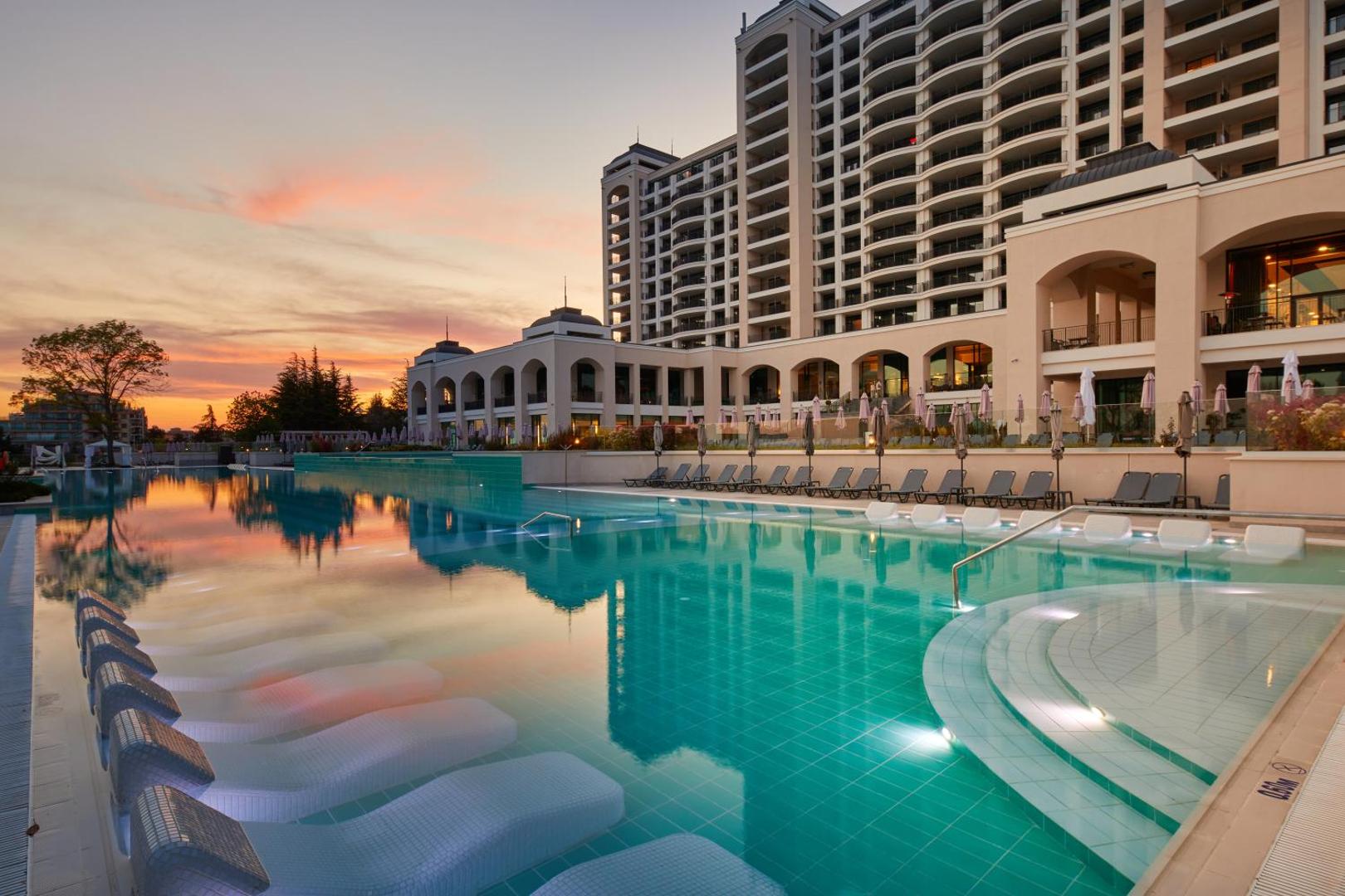 Secrets Sunny Beach Resort and Spa viesnīcas fasāde saulrietā ar apgaismotu āra baseinu un ūdens sauļošanās krēsliem