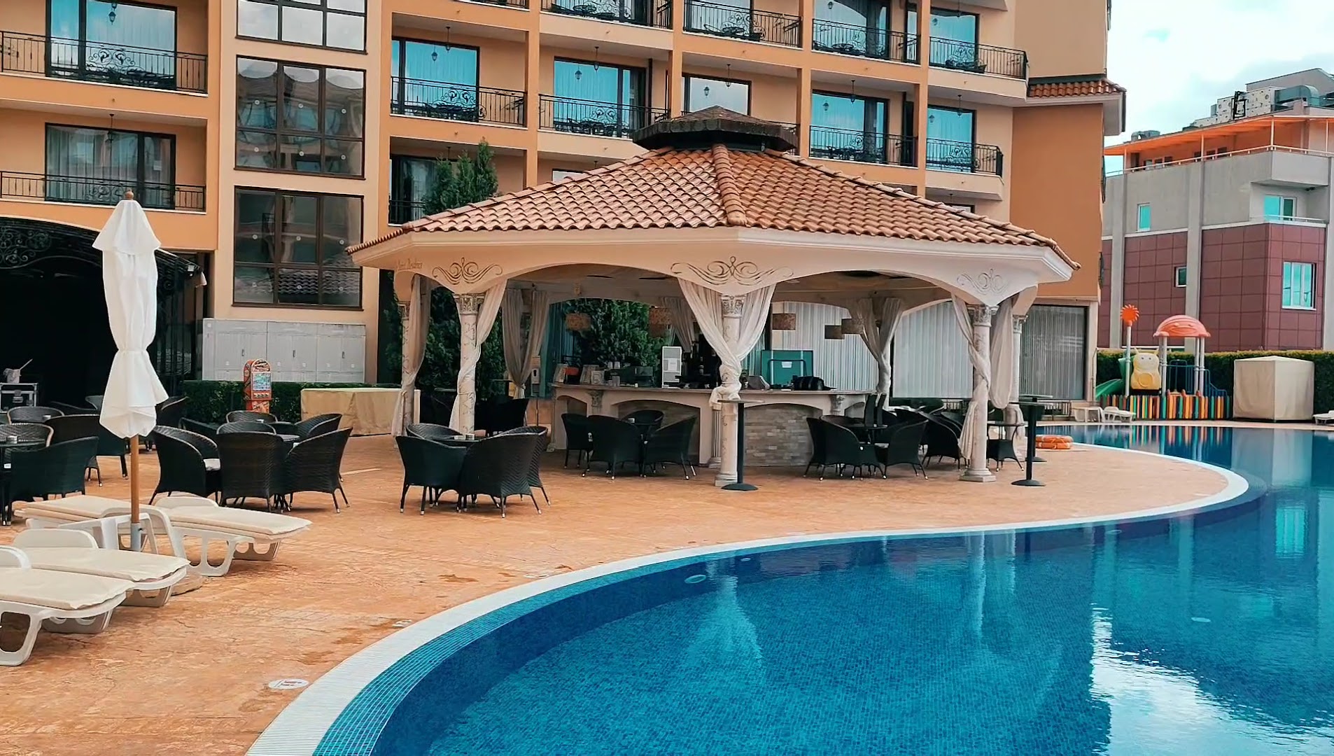 Diamant Residence Hotel & Spa baseina bārs ar nojumi un āra mēbelēm pie baseina malas, fonā bērnu rotaļu laukums