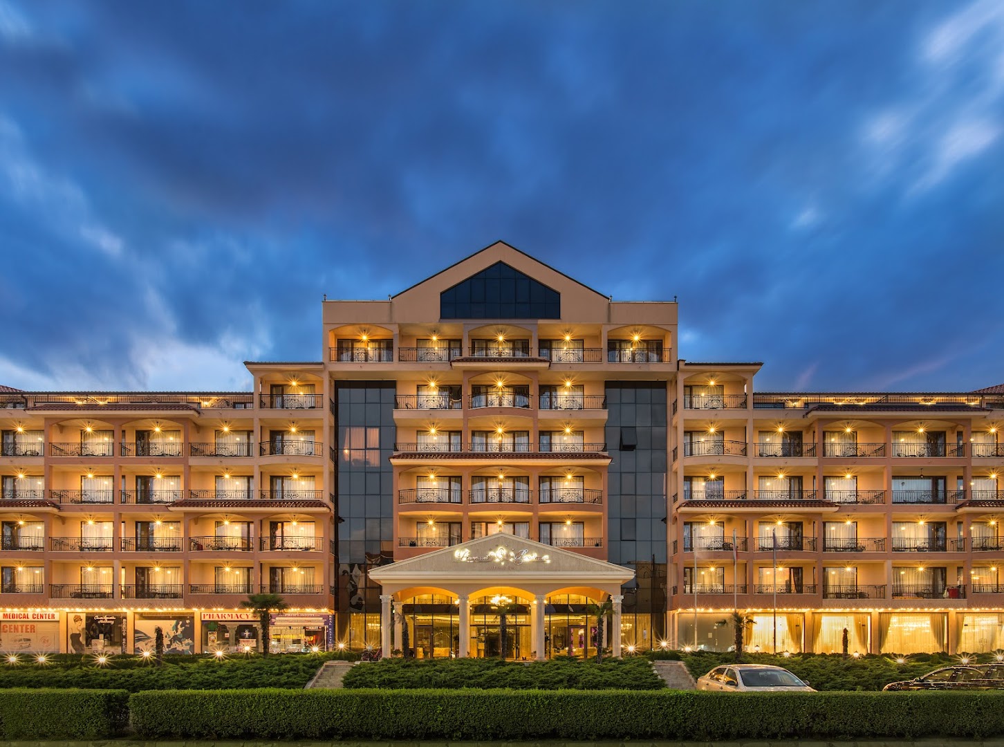 Diamant Residence Hotel & Spa fasāde vakarā Saulainajā krastā, Bulgārijā — apgaismoti balkoni un galvenā ieeja uz zila debesu fona