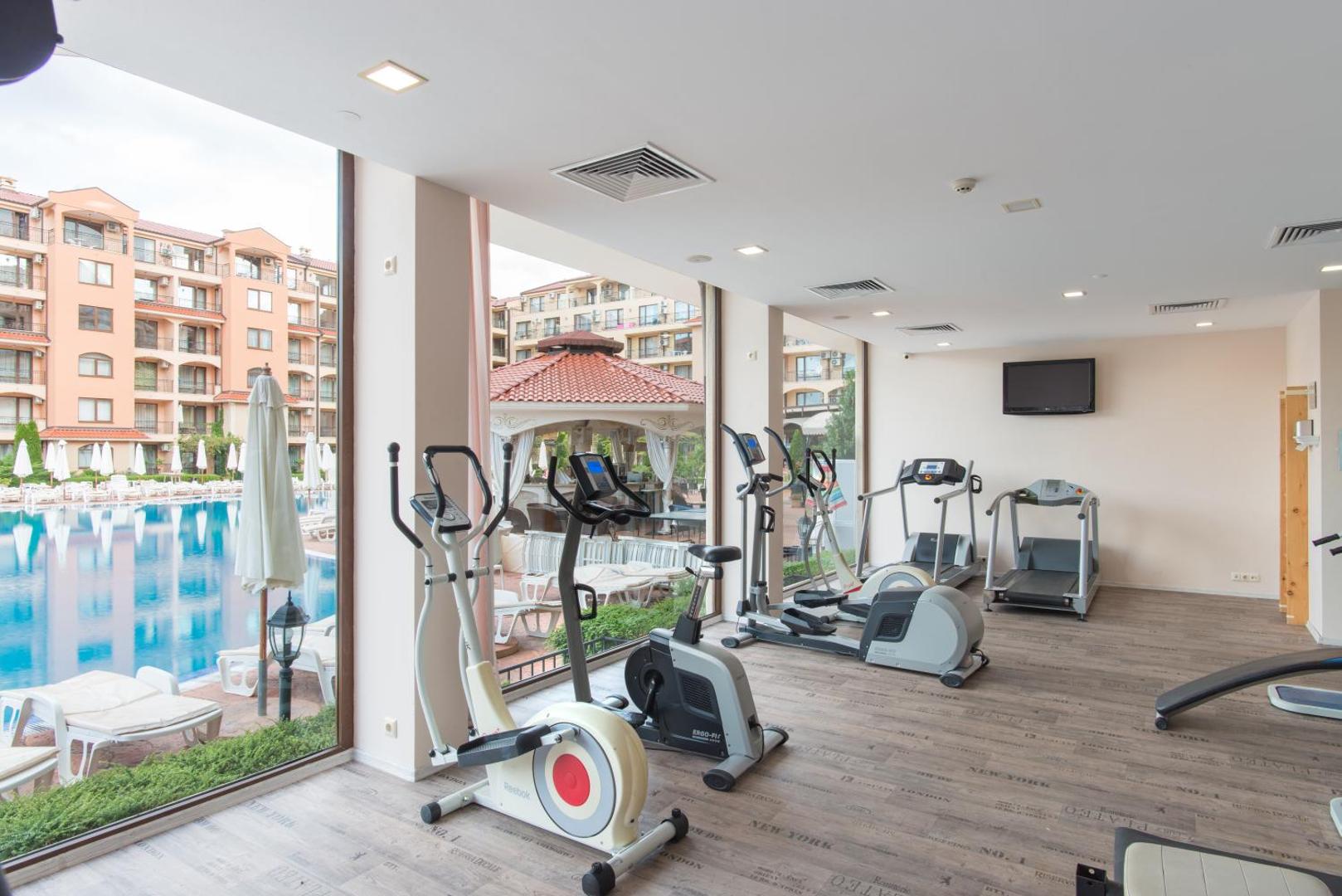 Diamant Residence Hotel & Spa fitness centrs ar skrejceļiem un velotrenažieriem, no kura paveras skats uz āra baseinu.