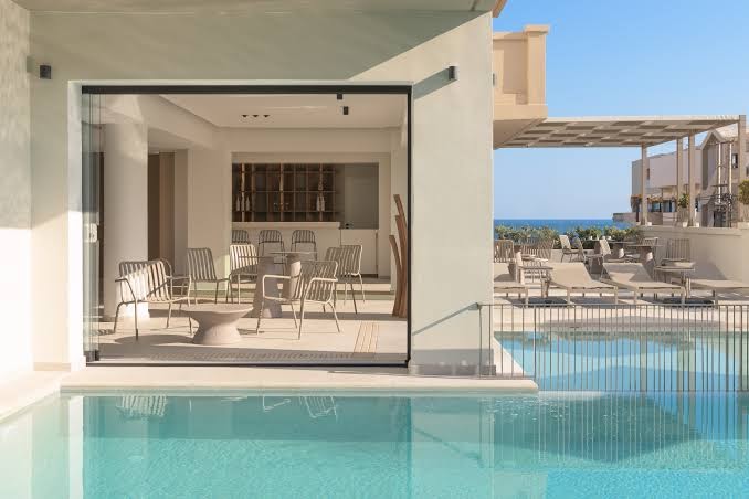 Theartemis Palace modernais bārs un atpūtas zona pie baseina — stikla sienas, dizaina mēbeles un skats uz jūru tālumā