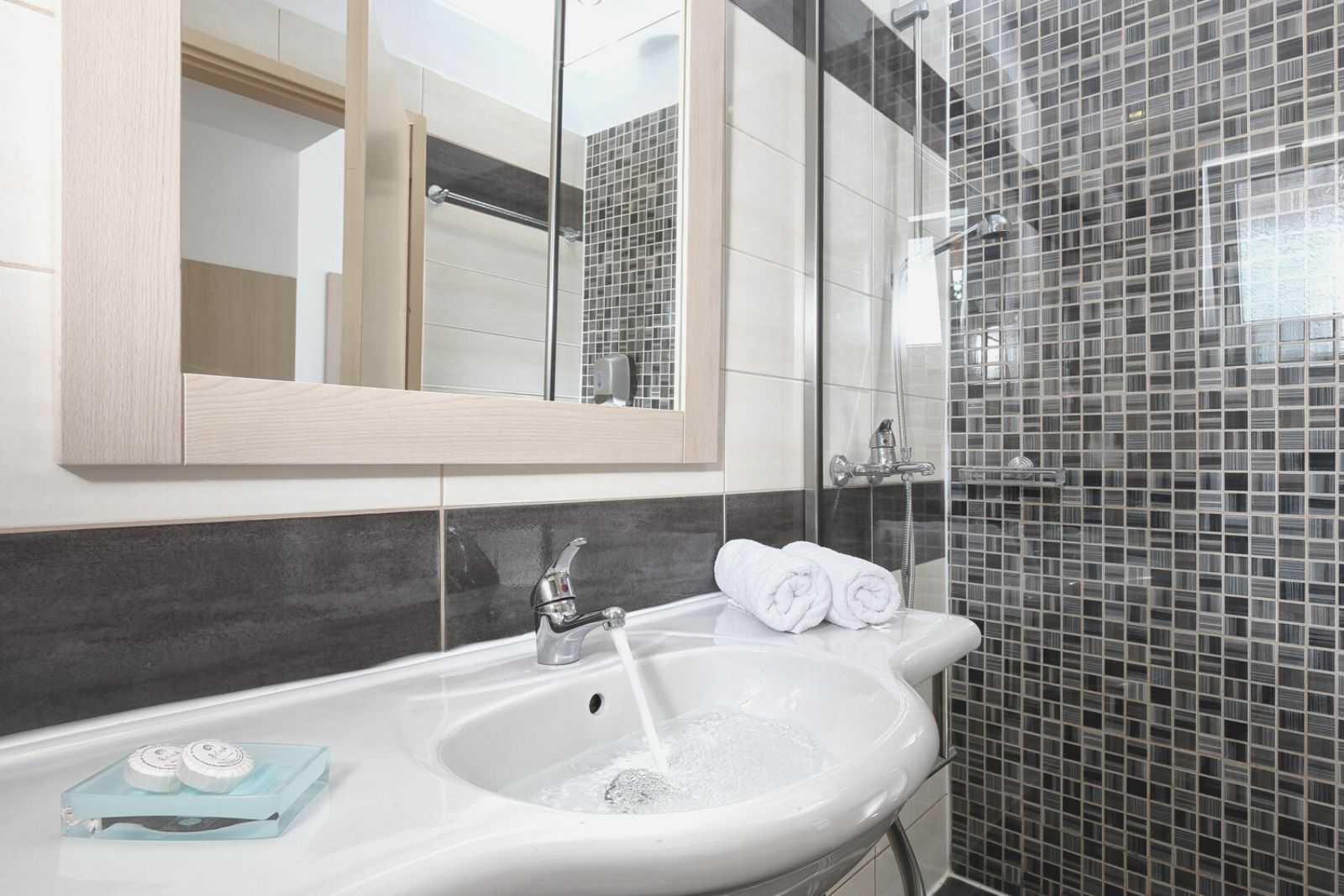 Vasia Hotel & Spa numura vannasistaba Sisī, Krētā — gaiša izlietne, spogulis koka rāmī un dušas zona ar pelēkām mozaīkas flīzēm