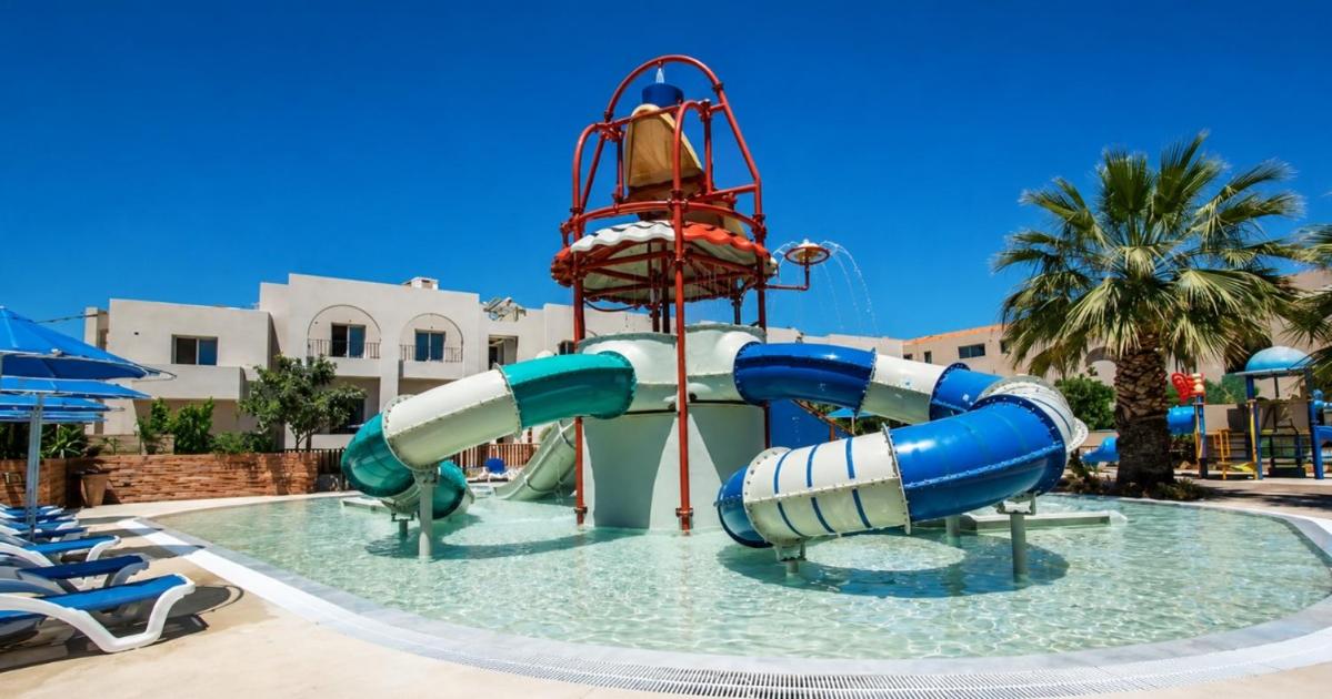 Vasia Hotel & Spa bērnu akvaparks ar krāsainiem slidkalniņiem un seklu baseinu, fonā redzamas viesnīcas ēkas un palmas