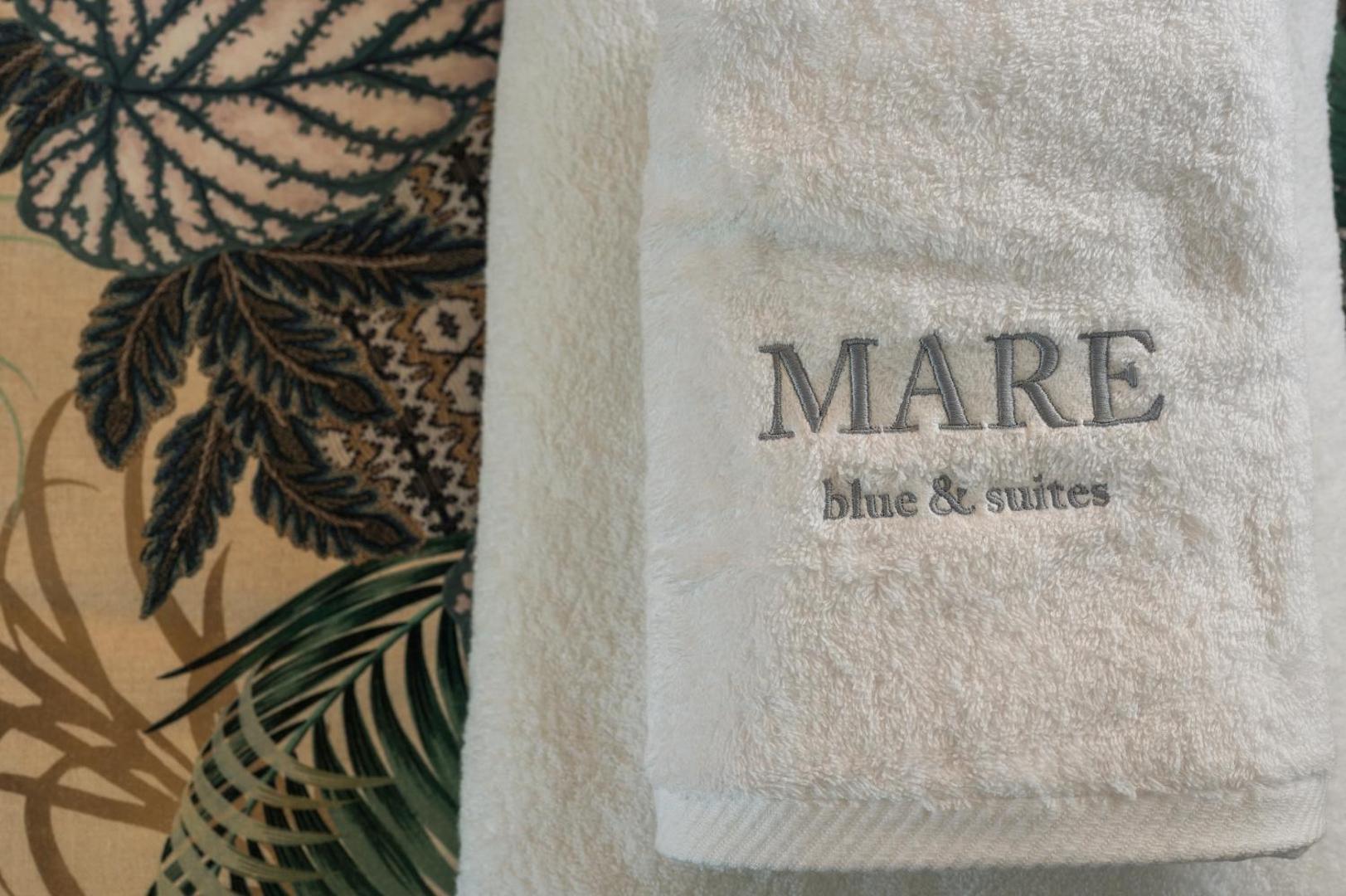 Mīksti balti dvieļi ar Mare Blue Suites izšūtu logo uz dekoratīva tropu raksta fona viesnīcas numurā
