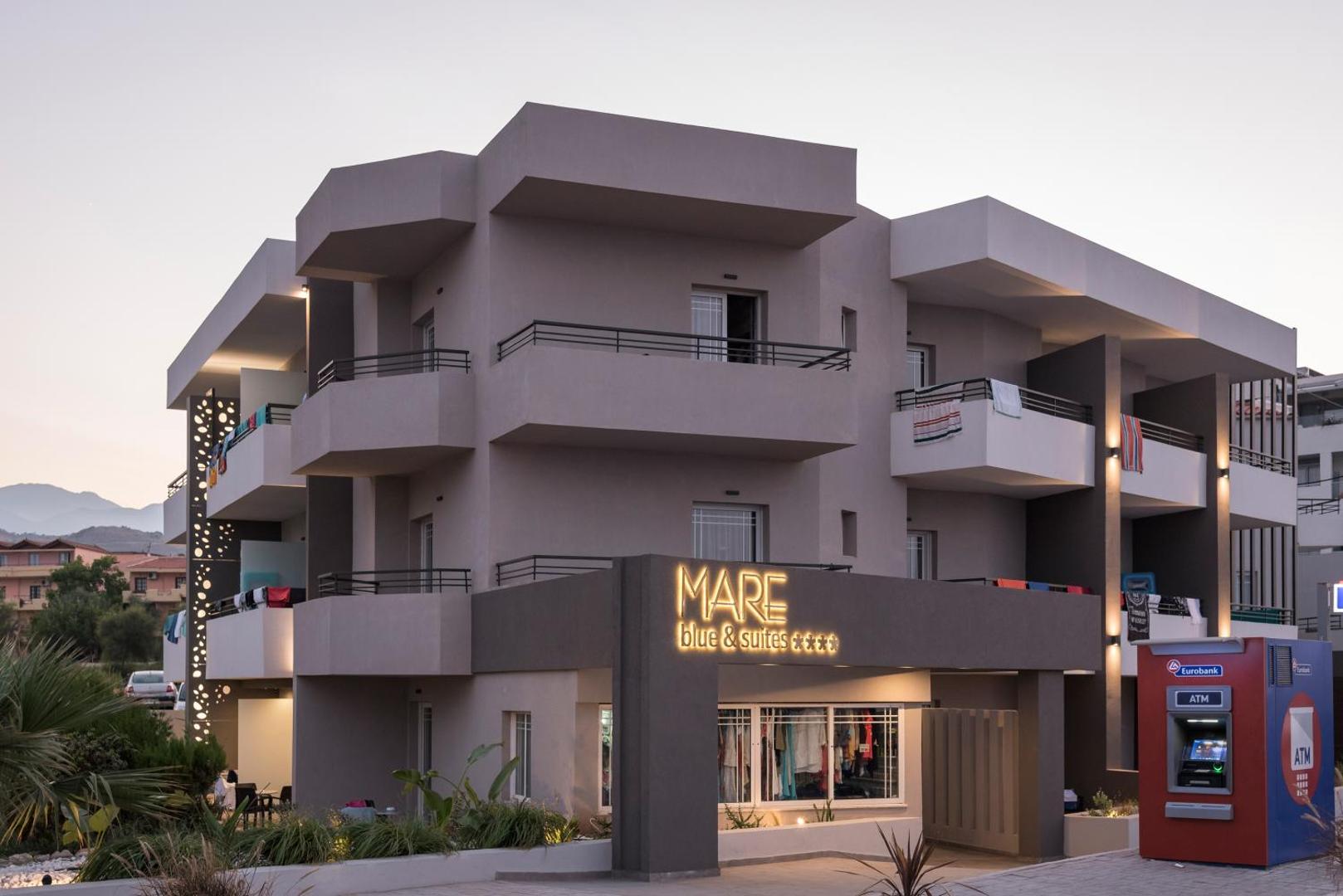 Mare Blue Suites viesnīcas fasāde vakarā Krētā — moderna četru stāvu ēka ar balkoniem un izgaismotu logo pie ieejas.