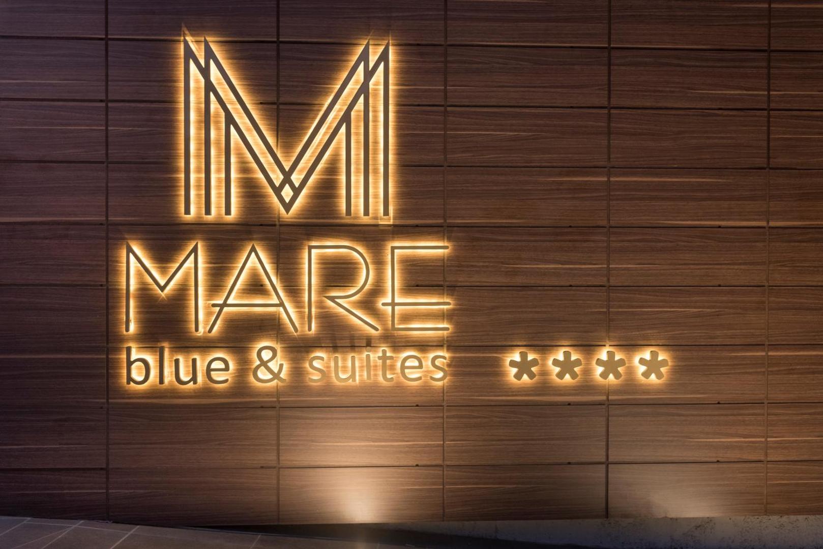 Mare Blue Suites 4* viesnīcas izgaismotais logo uz koka apdares sienas pie ieejas vakarā