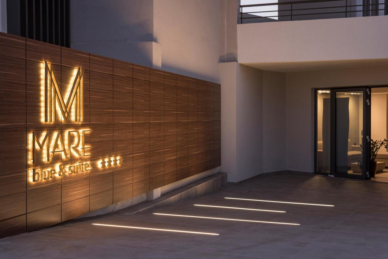 Mare Blue Suites 4* viesnīcas ieeja ar izgaismotu koka paneļu sienu un zīmola logo piekrastes kūrortā Krētā.
