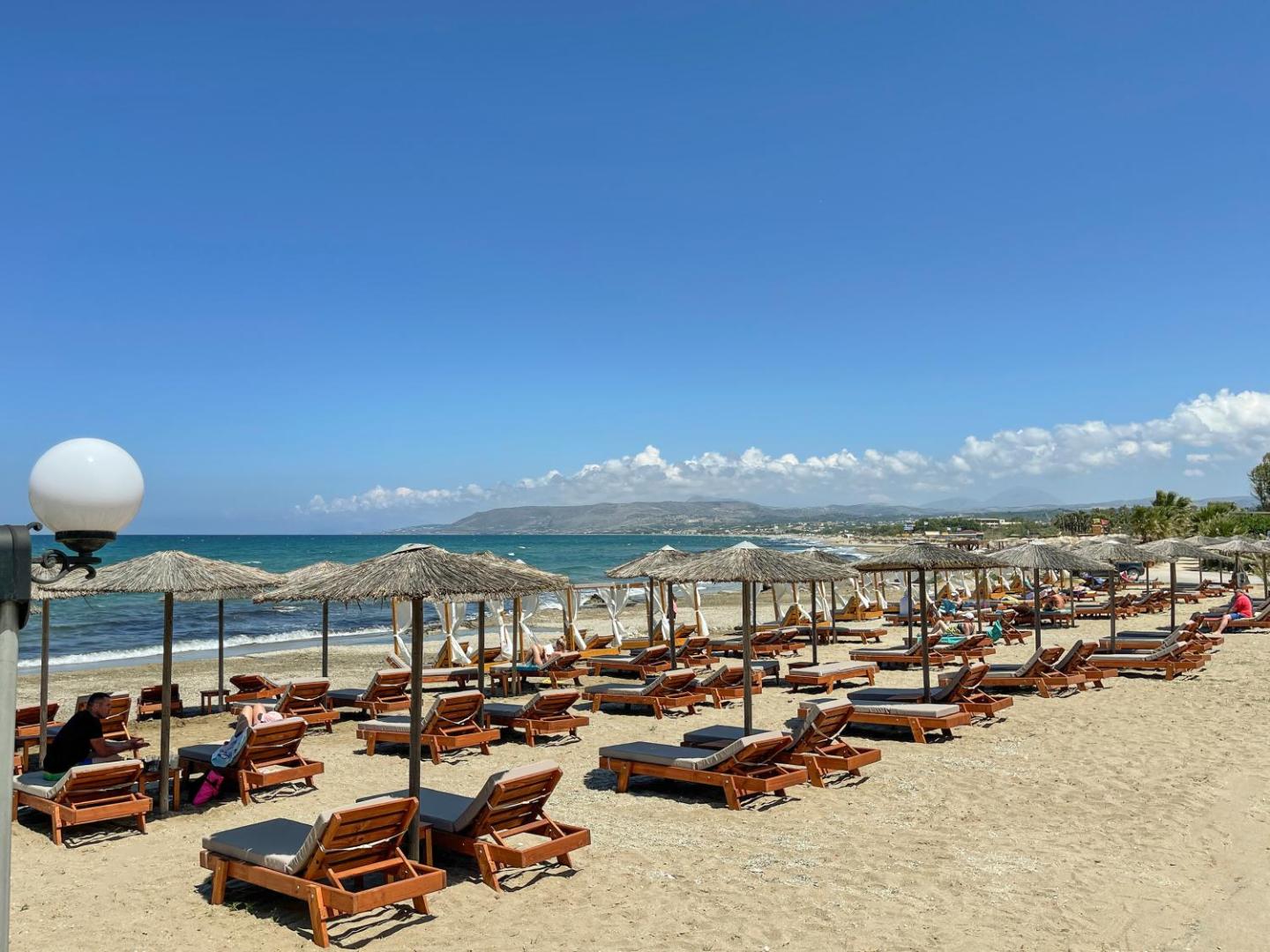 Mare Blue Suites privātā smilšu pludmale ar koka sauļošanās krēsliem un salmu saulessargiem pie zilās jūras Krētas piekrastē