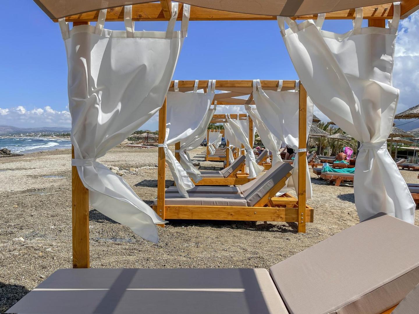 Mare Blue Suites pludmales atpūtas zona ar koka bungalo tipa saulessargiem un mīkstiem zviļņiem Krētas piekrastē, Stalidā.