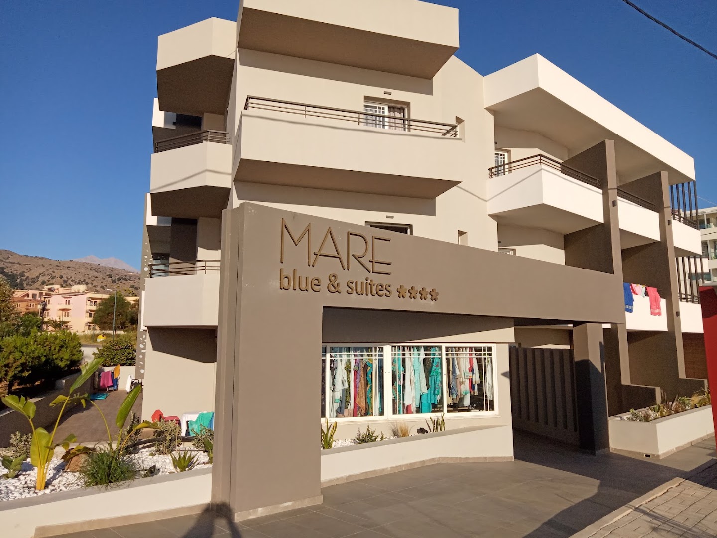 Mare Blue Suites viesnīcas ēka dienas gaismā ar balkoniem un kalnu fonu Krētas salā, Grieķijā.