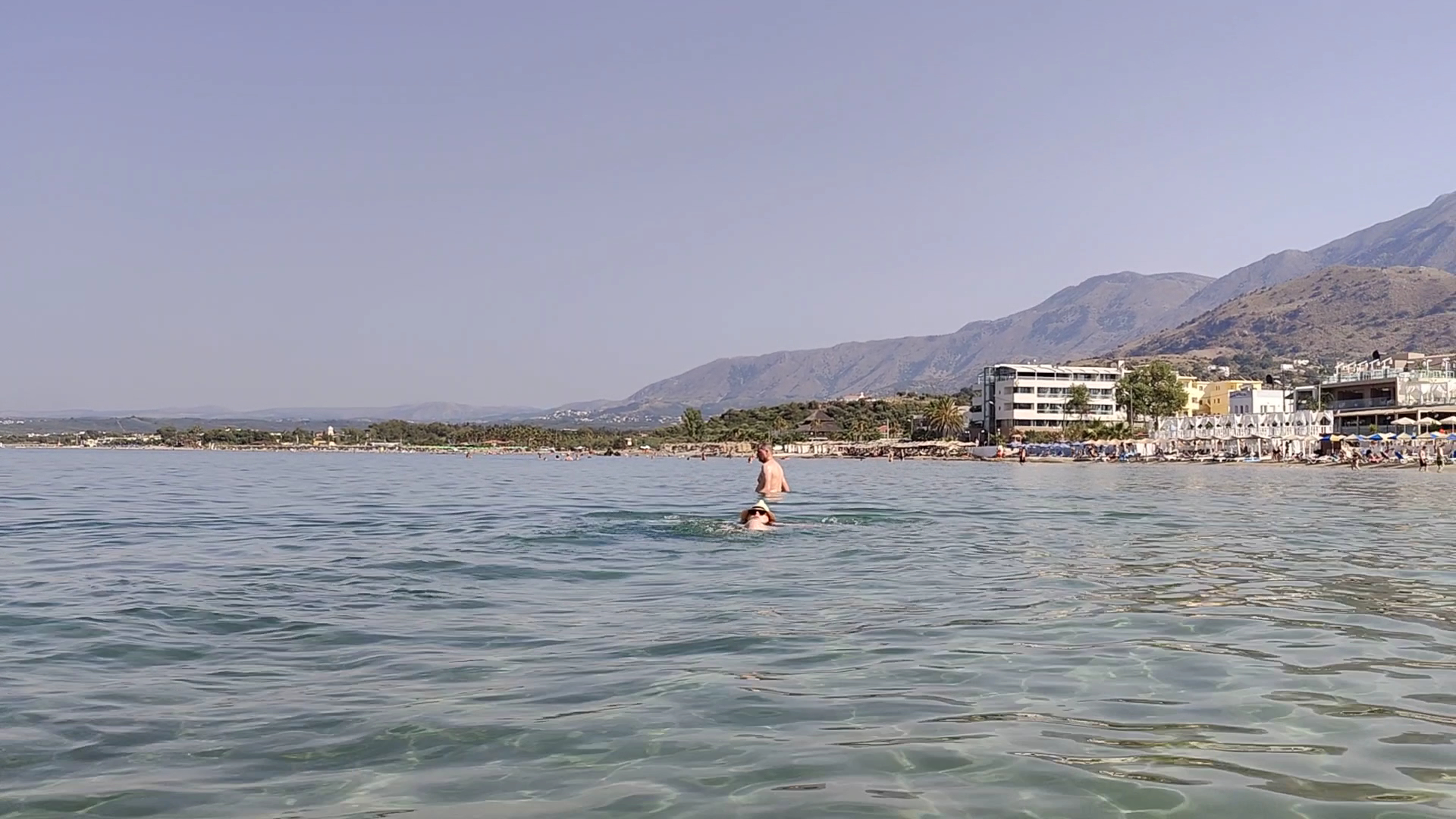 Corissia Beach Hotel skats no jūras uz Georgiupoles pludmali Krētā — tirkīzzils ūdens, kalnu fons un viesnīcas ēkas piekrastē