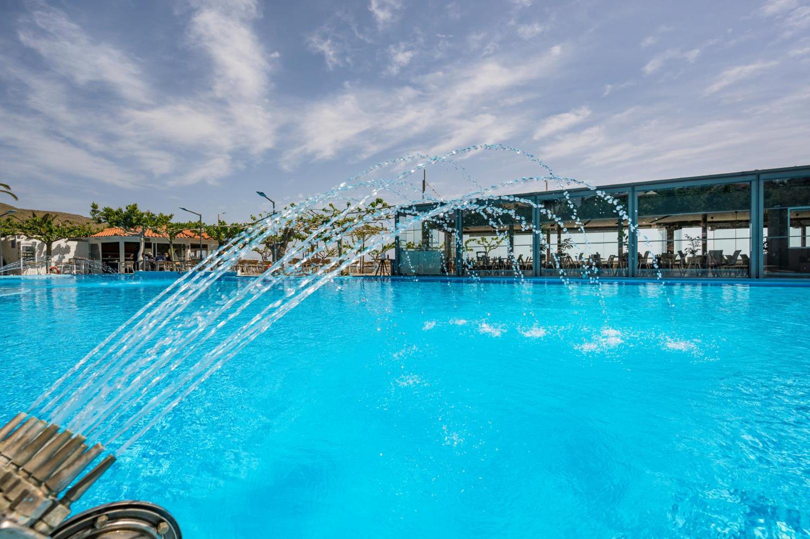 Corissia Beach Hotel āra peldbaseins ar dekoratīvām ūdens strūklakām Georgiupoli, Krētā — fona redzama restorāna terase un zilas debesis