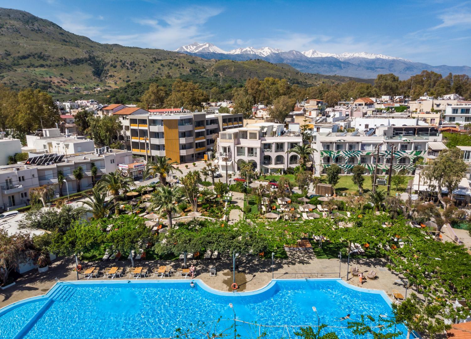 Corissia Beach Hotel aerofoto ar lielu āra baseinu, atpūtas zonu un skatu uz sniegotajām Krētas kalnu virsotnēm tālumā.