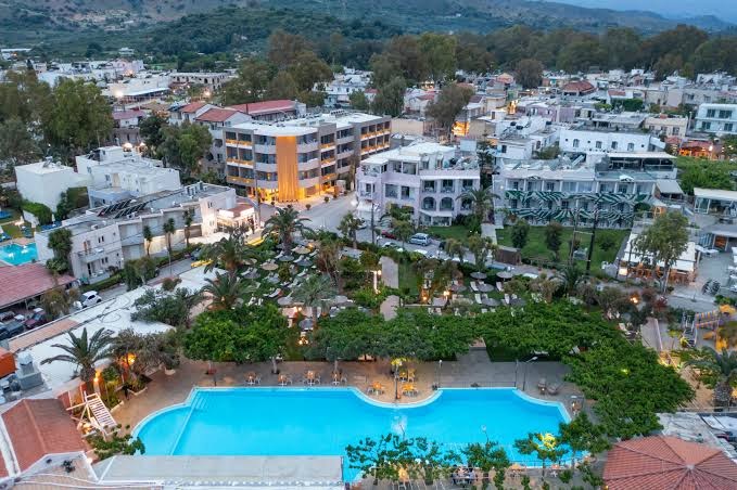 Corissia Beach Hotel teritorijas skats no putna lidojuma ar plašu āra baseinu, atpūtas zonu un apkārtējo pilsētas ainavu Krētā.