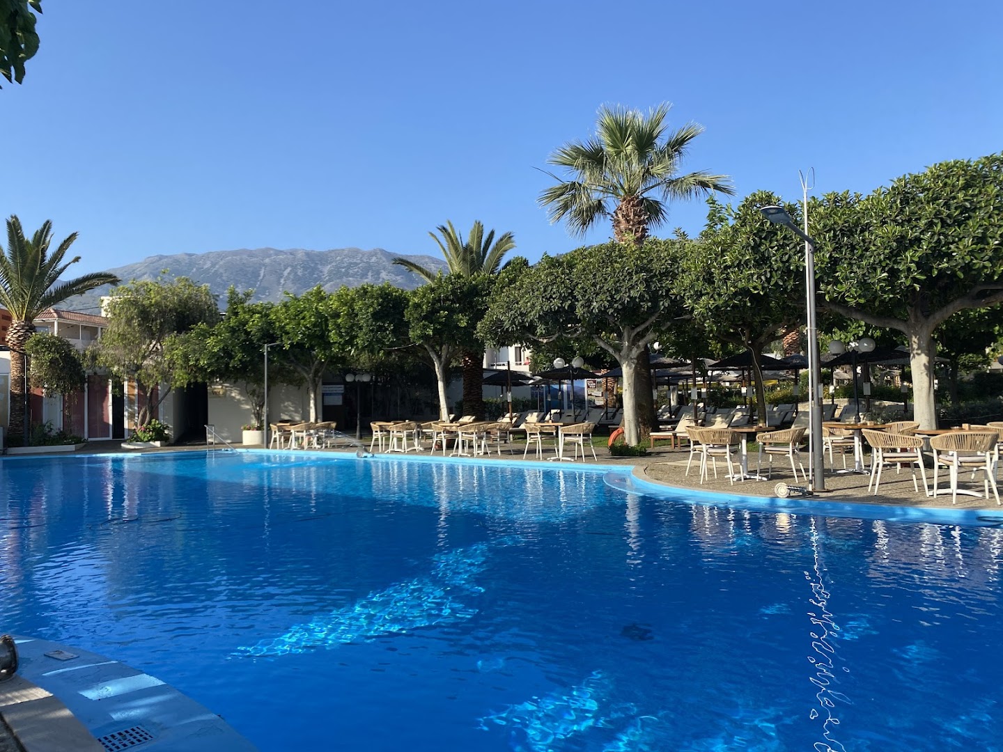 Corissia Beach Hotel plašais āra baseins Georgiupoli kūrortā — apkārtnē kopti dārzi, palmas, atpūtas krēsli un kalnu ainava fonā