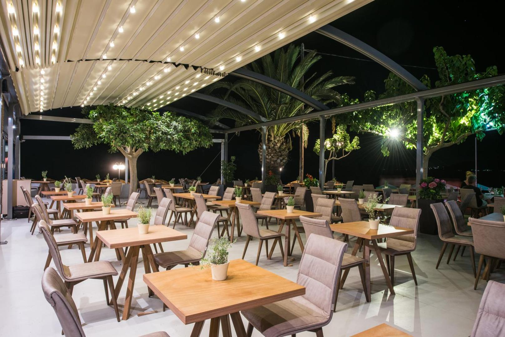 Corissia Beach Hotel restorāna āra terase vakarā ar modernu apgaismojumu un koka galdiem Georgiupoli kūrortā, Krētā
