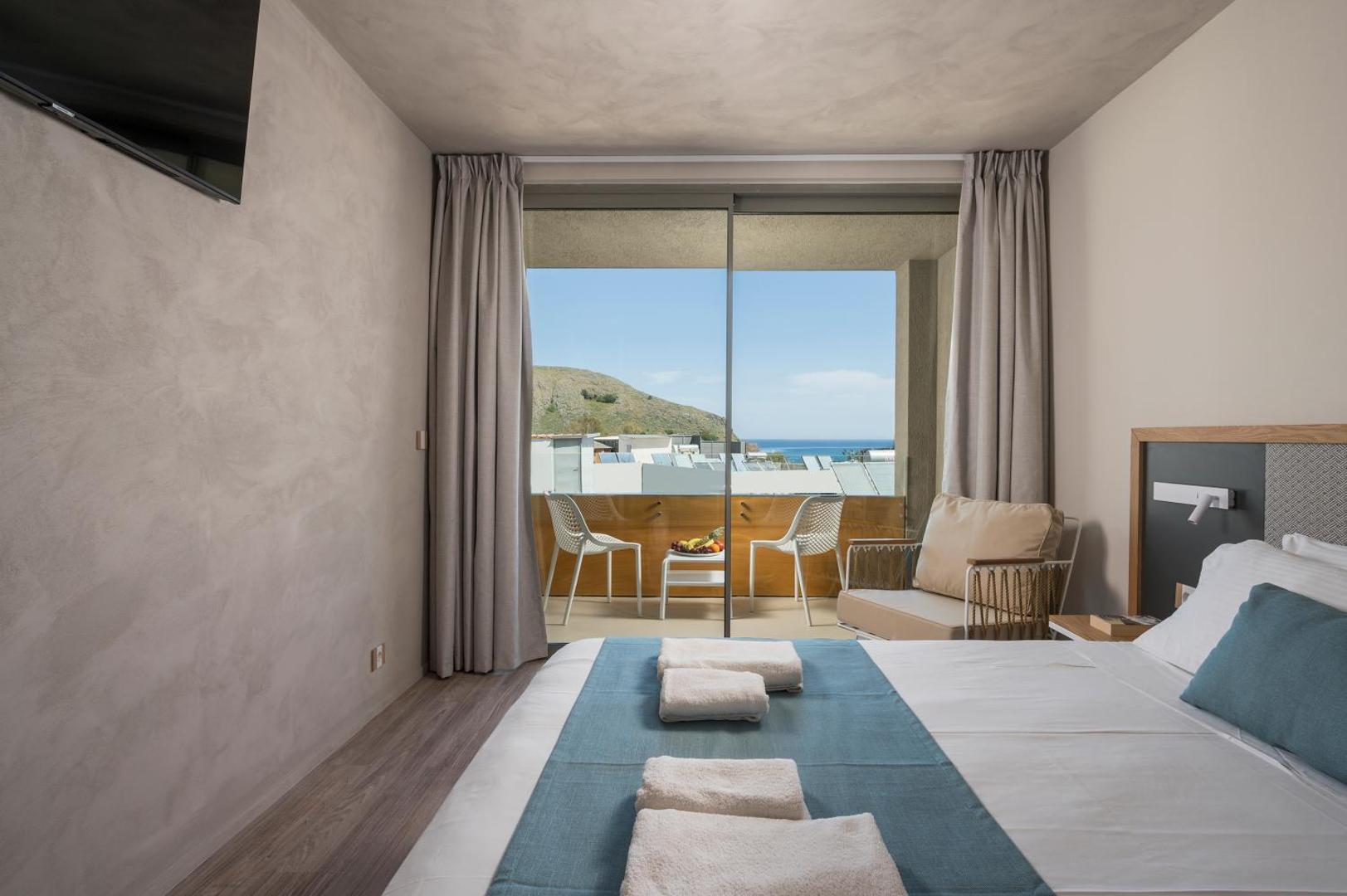 Corissia Beach Hotel gaišs numurs ar balkonu, no kura paveras skats uz jūru un kalniem — moderna iekārtojuma interjers.