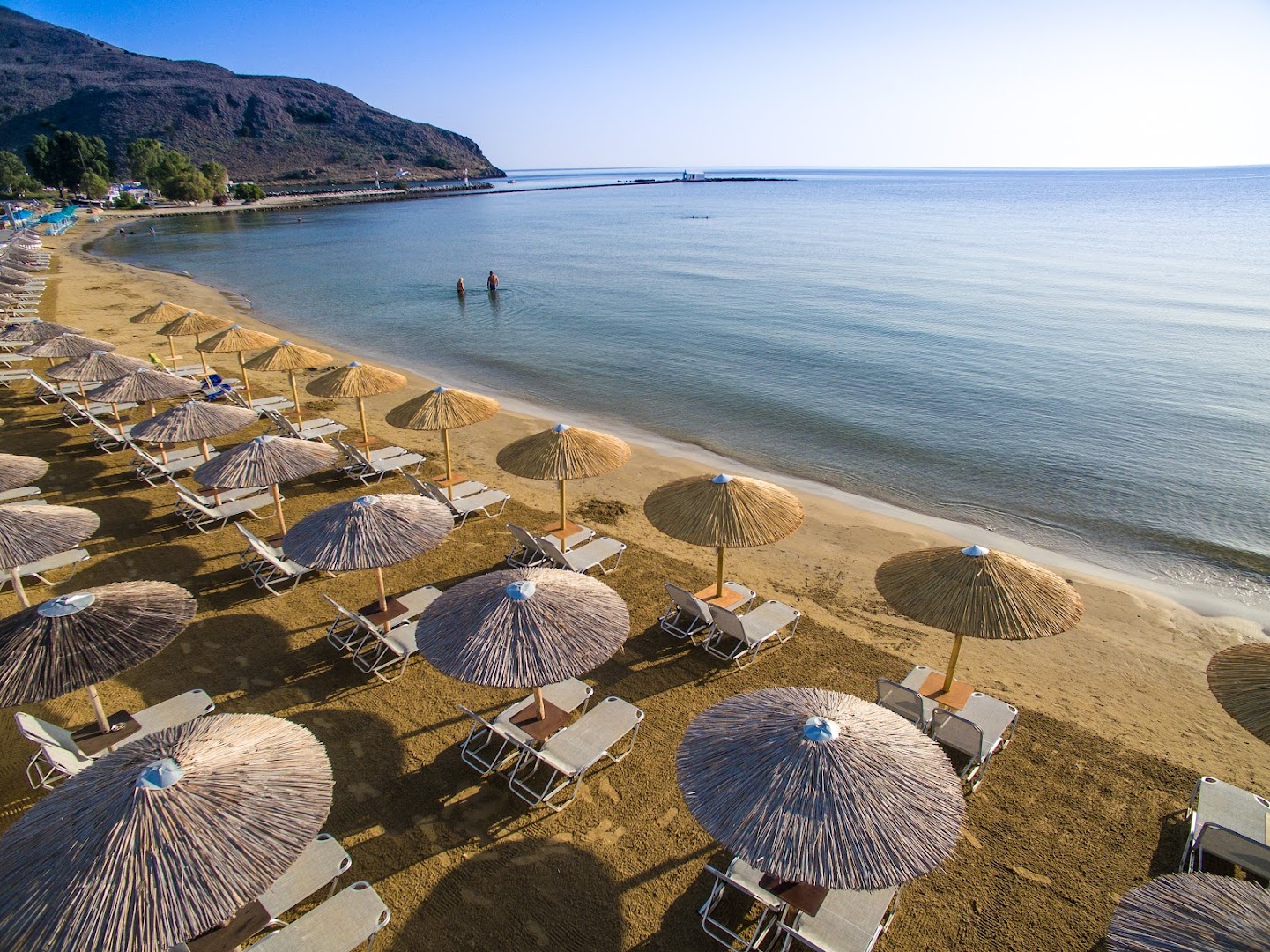Corissia Beach Hotel privātā smilšu pludmale ar saulessargiem un zviļņiem Georgiupoli līcī, Krētā — skats uz mierīgu jūru un kalniem