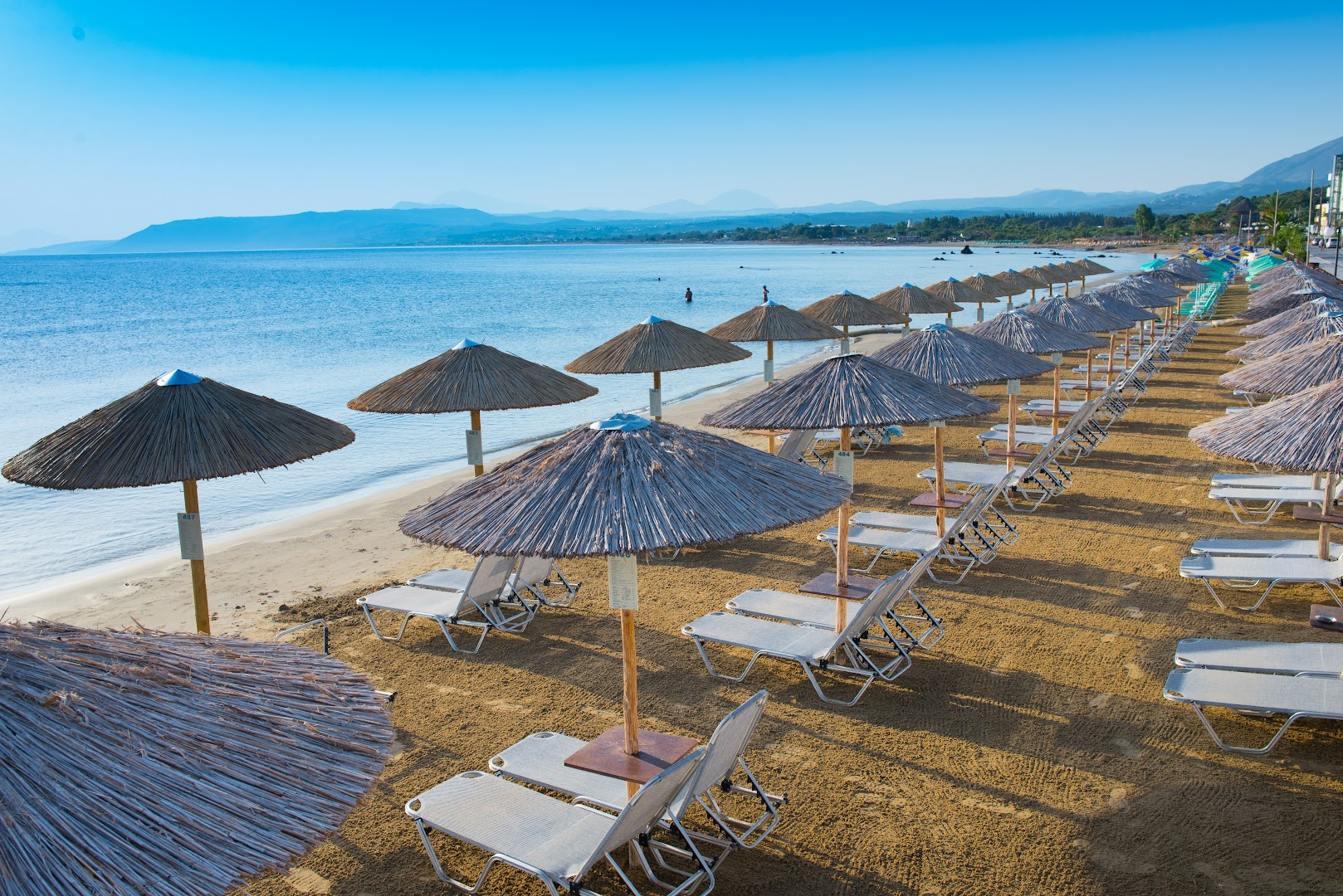 Skats uz Corissia Beach Hotel pludmales zonu ar sakārtotām sauļošanās vietām un salmu saulessargiem pie dzidrās Vidusjūras Georgiupoli