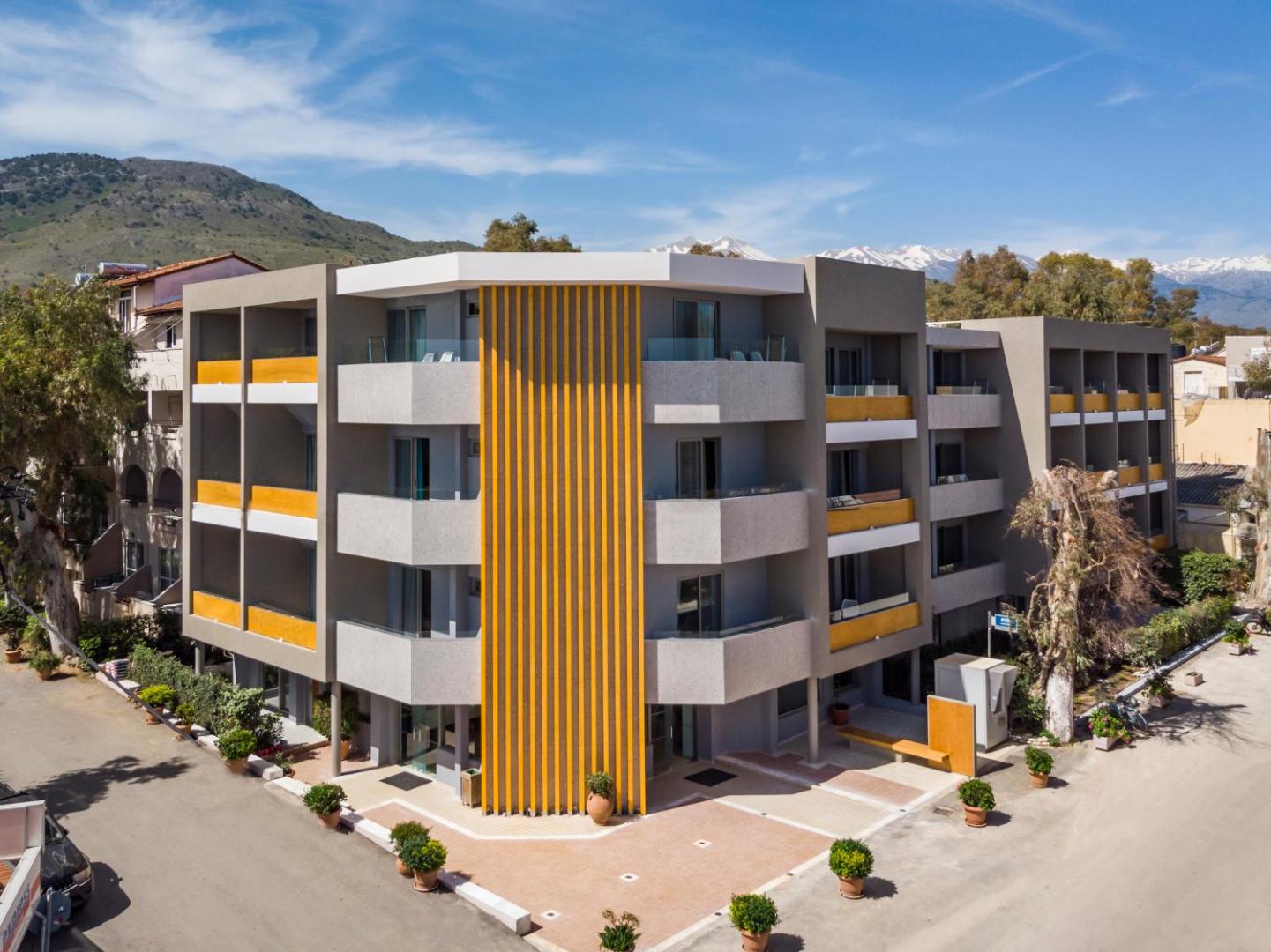 Corissia Beach Hotel viesnīcas ēka dienas gaismā ar skatu uz sniegotajām kalnu virsotnēm tālumā un sakoptu apkārtni.