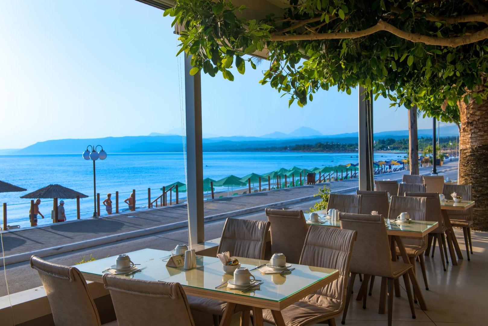 Corissia Beach Hotel restorāna āra terase ar skatu uz jūru un pludmali — galdiņi servēti brokastīm zem koku lapotnes