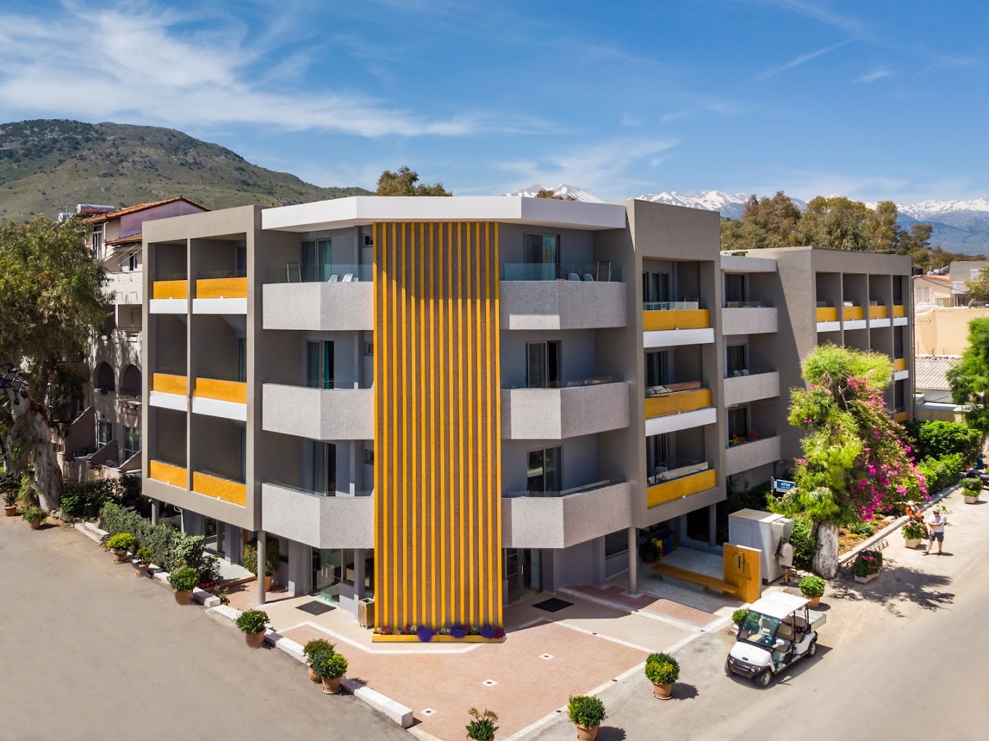 Corissia Beach Hotel modernā fasāde Georgiupoli, Krētā — skats no augšas uz četru stāvu ēku ar balkoniem un kalnu fonu.