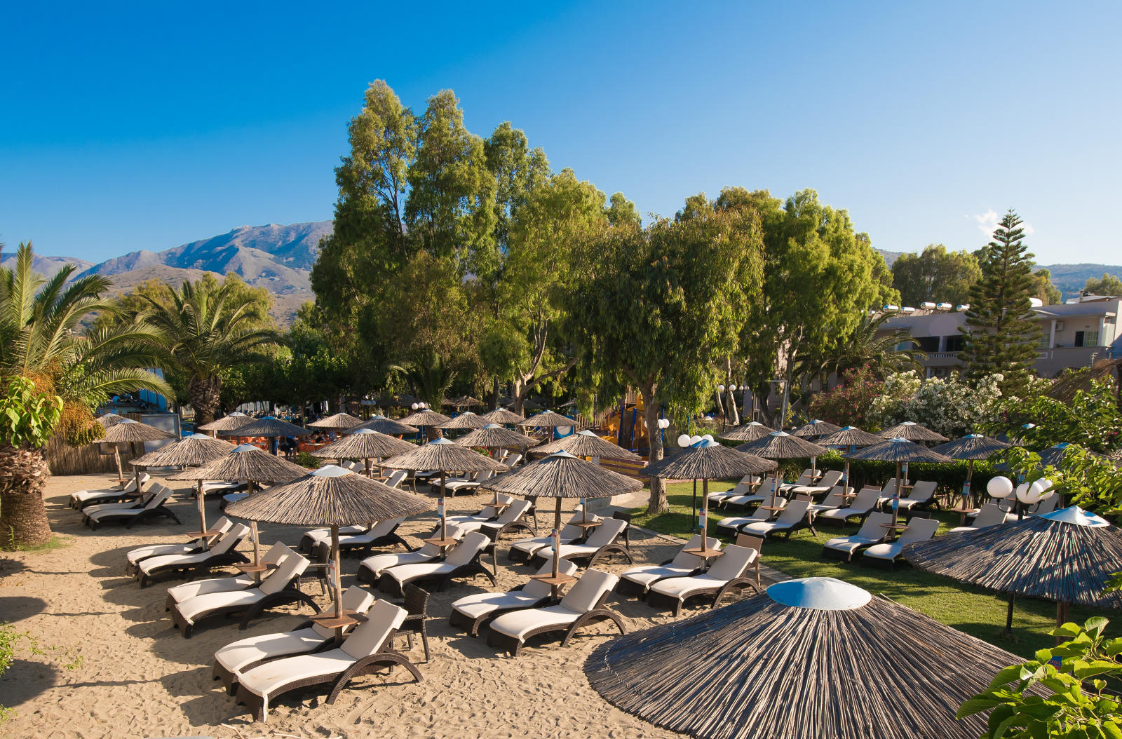 Corissia Beach Hotel atpūtas zona ar zviļņiem un saulessargiem dārza un pludmales pierobežā, fonā redzami Krētas kalni