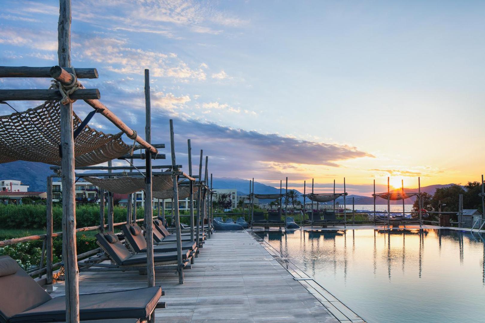 La Mer Resort & Spa baseina zona saulrietā ar koka sauļošanās krēsliem un dekoratīvām nojumēm Krētas piekrastē.