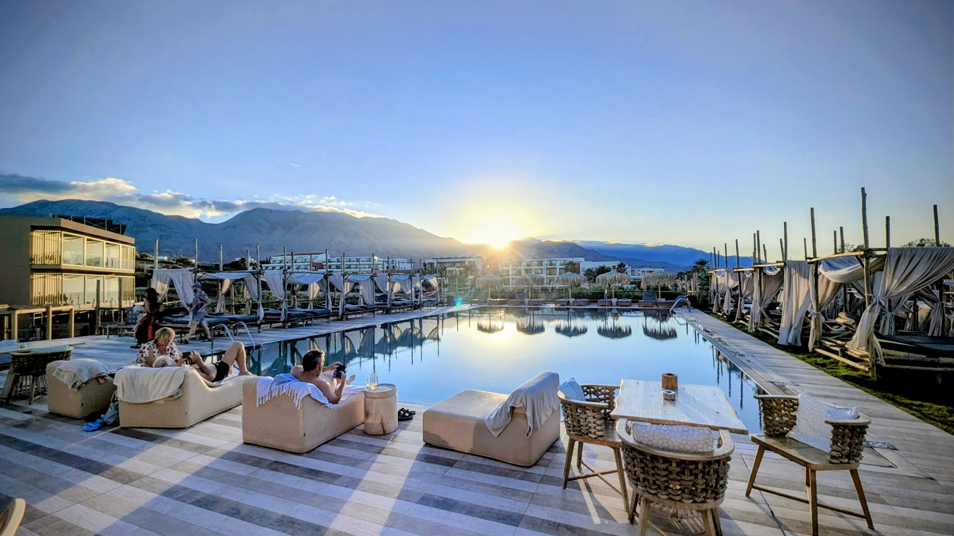 La Mer Resort & Spa plašā baseina zona ar atpūtas krēsliem, galdiņiem un skatu uz kalniem saulrietā Krētā.