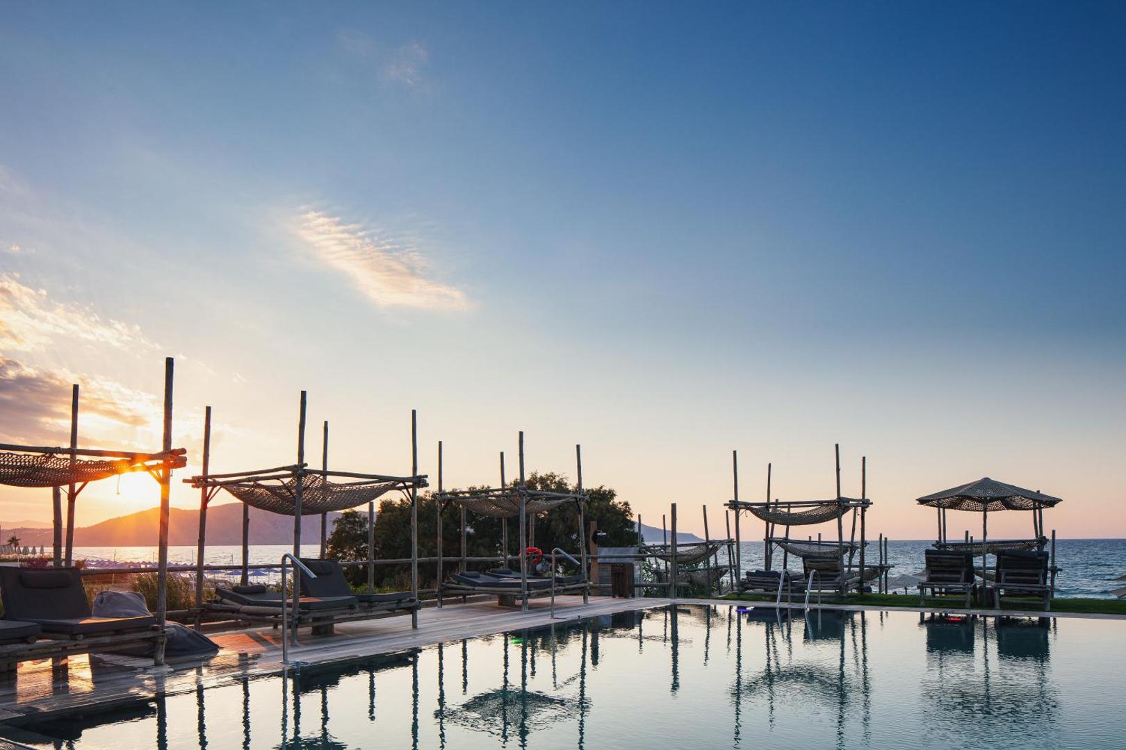 La Mer Resort & Spa baseins ar skatu uz jūru un kalniem saulrieta laikā, koka konstrukcijas un atpūtas krēsli.