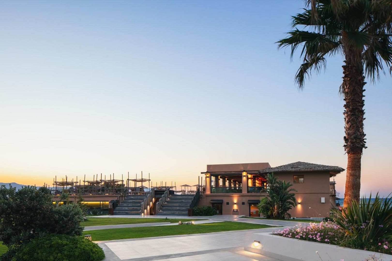La Mer Resort & Spa viesnīcas teritorija saulrietā ar restorāna ēku, dārzu un palmām Krētas piekrastē