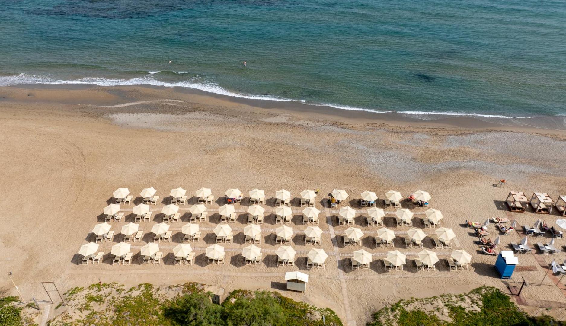Orpheas Resort Hotel privātā pludmale ar gaišiem saulessargiem un sauļošanās krēsliem pie dzidri zilās jūras Krētas salā