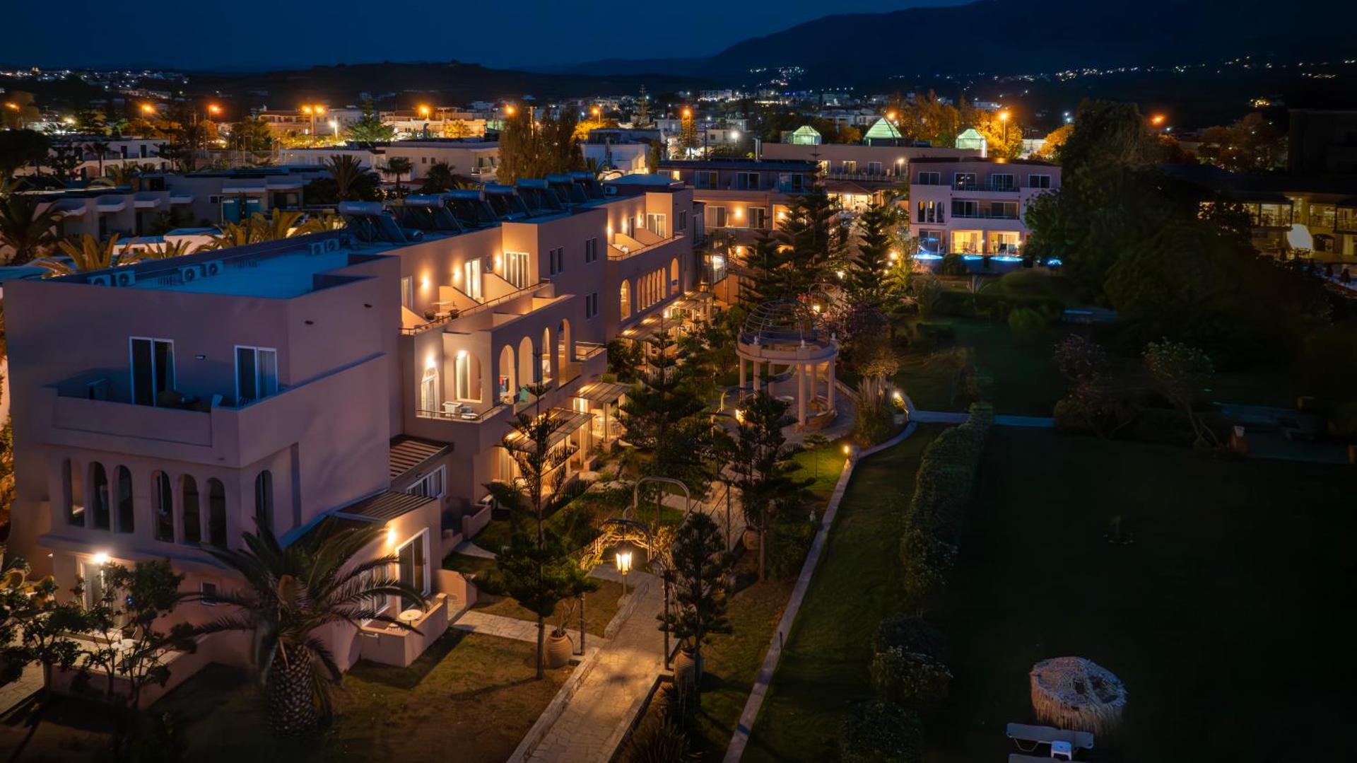 Orpheas Resort Hotel ēku fasādes un dārzs vakara apgaismojumā ar izgaismotiem balkoniem un gājēju celiņiem