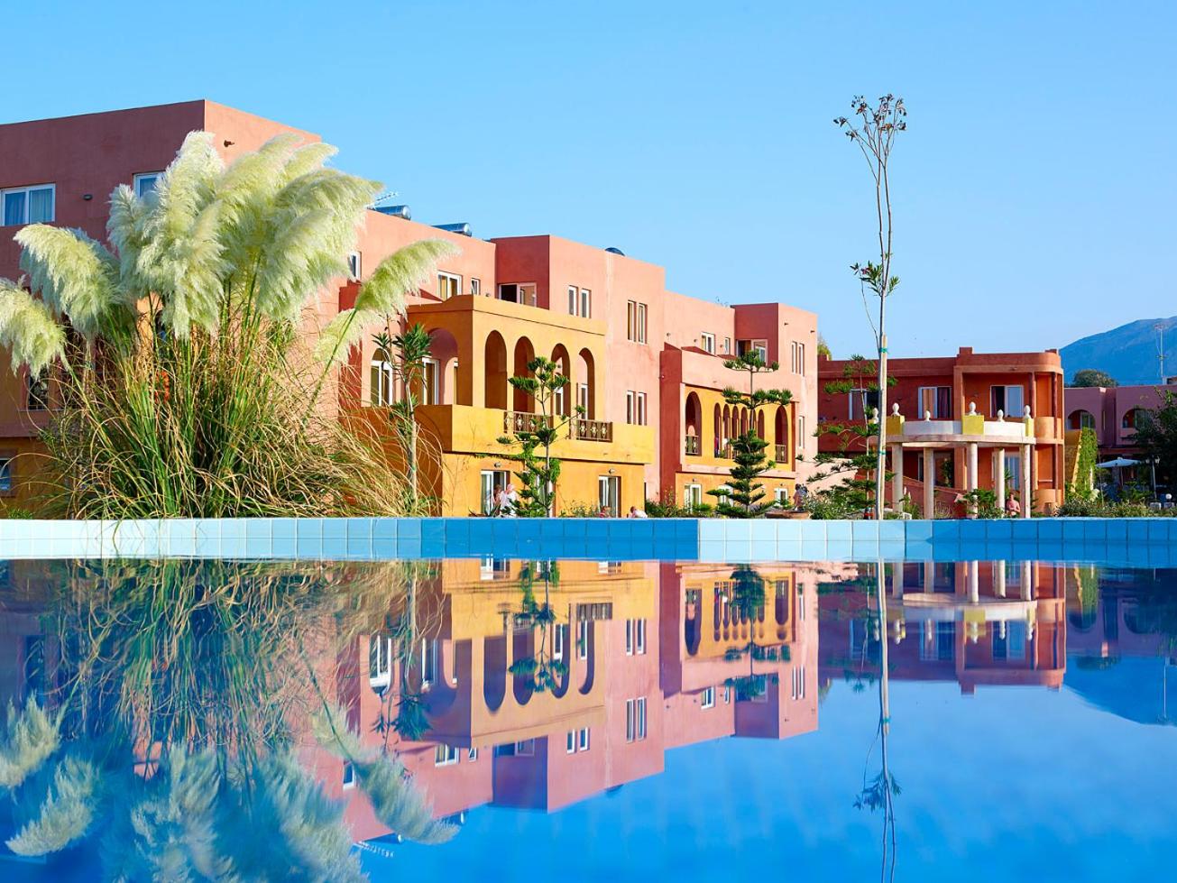 Orpheas Resort Hotel ēkas atspulgs dzidrā baseina ūdenī, fonā terakotas krāsas numuru korpusi