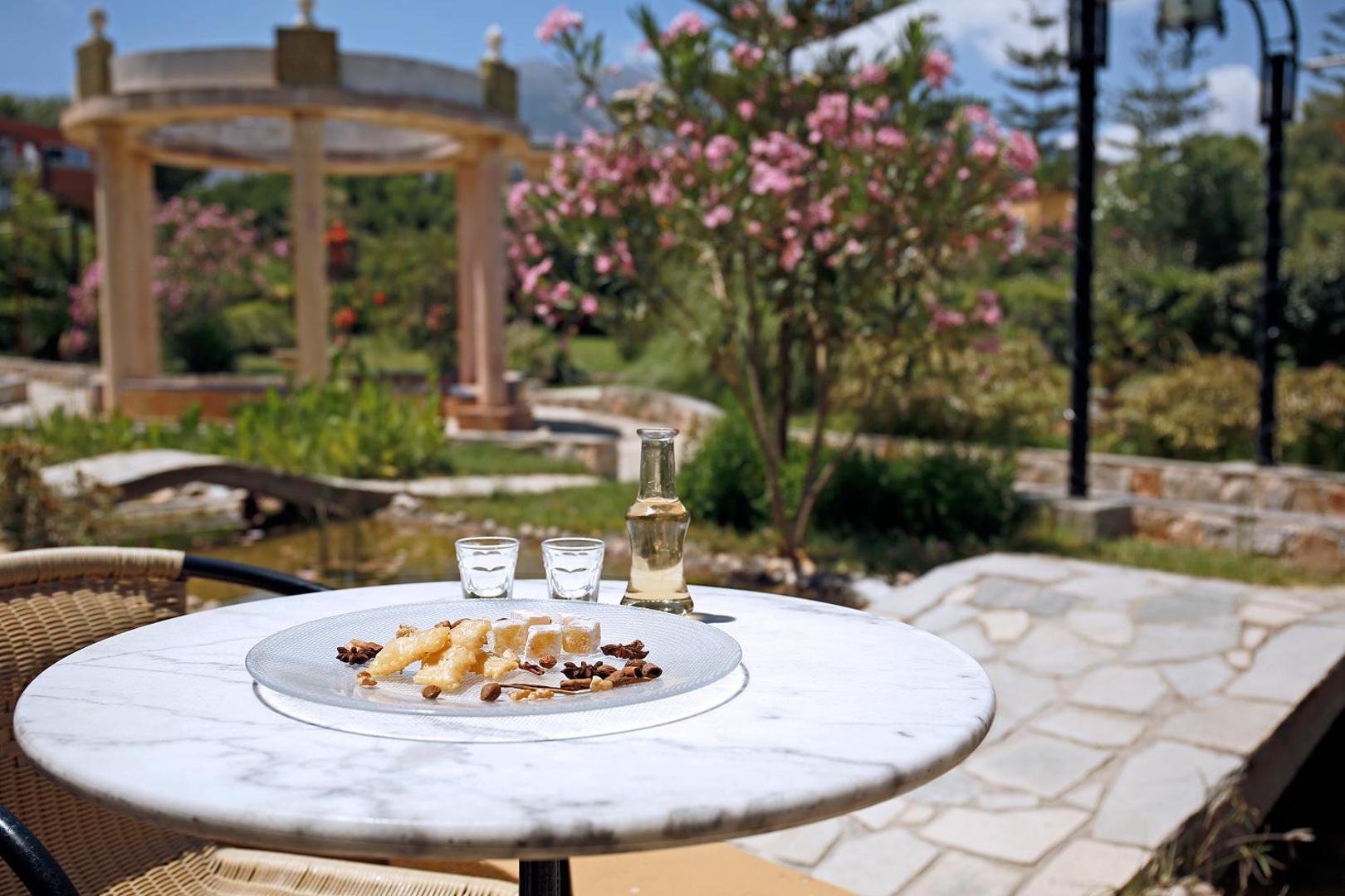 Orpheas Resort Hotel dārzā uz galda servēti grieķu saldumi un raki dzēriens, fonā ziedoši krūmi un dekoratīva lapene Krētā